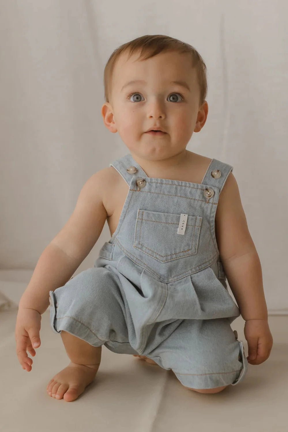 A baby with short brown hair and blue eyes is dressed in ethically made Denim Short Overalls Baby Blue by SUSUKOSHI, crafted from organic cotton, over a diaper. The baby is kneeling on a cream-colored floor, gazing upward with a curious expression. The background is a neutral shade.