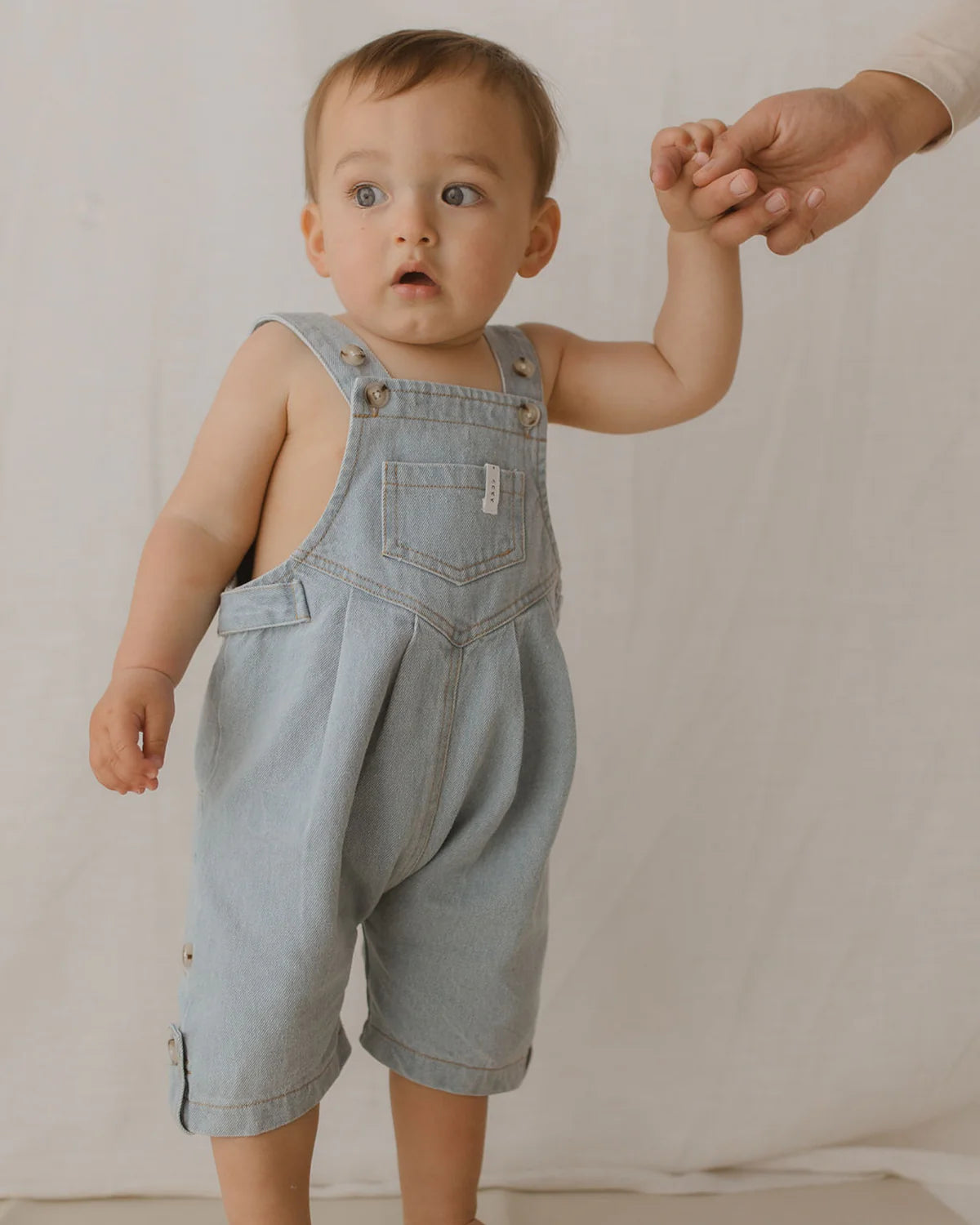 A toddler wearing Denim Short Overalls in Baby Blue by SUSUKOSHI, ethically made in China from organic cotton, stands while holding an adult's hand. The child has short hair and a focused expression, with a simple and neutral background emphasizing the child as the main subject.