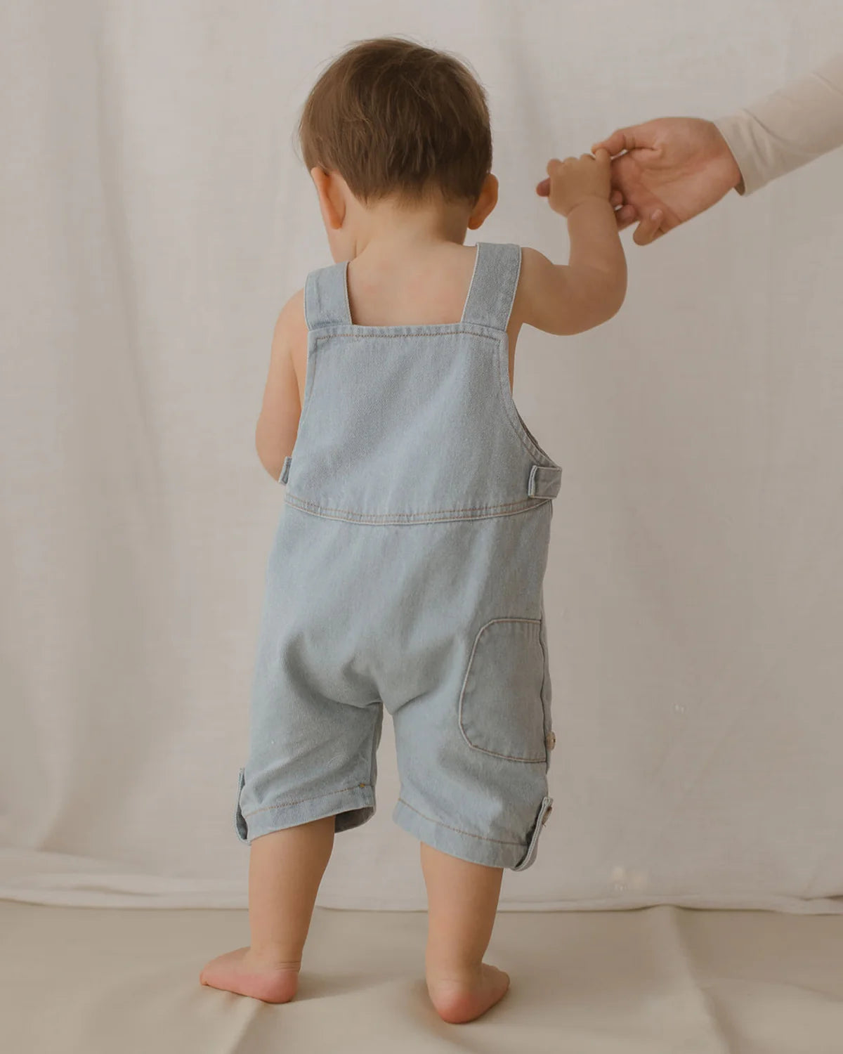 A child in ethically made Denim Short Overalls Baby Blue by SUSUKOSHI, seen from the back, holds an adult's hand. The child is barefoot and standing on a soft, light-colored surface with a neutral background.