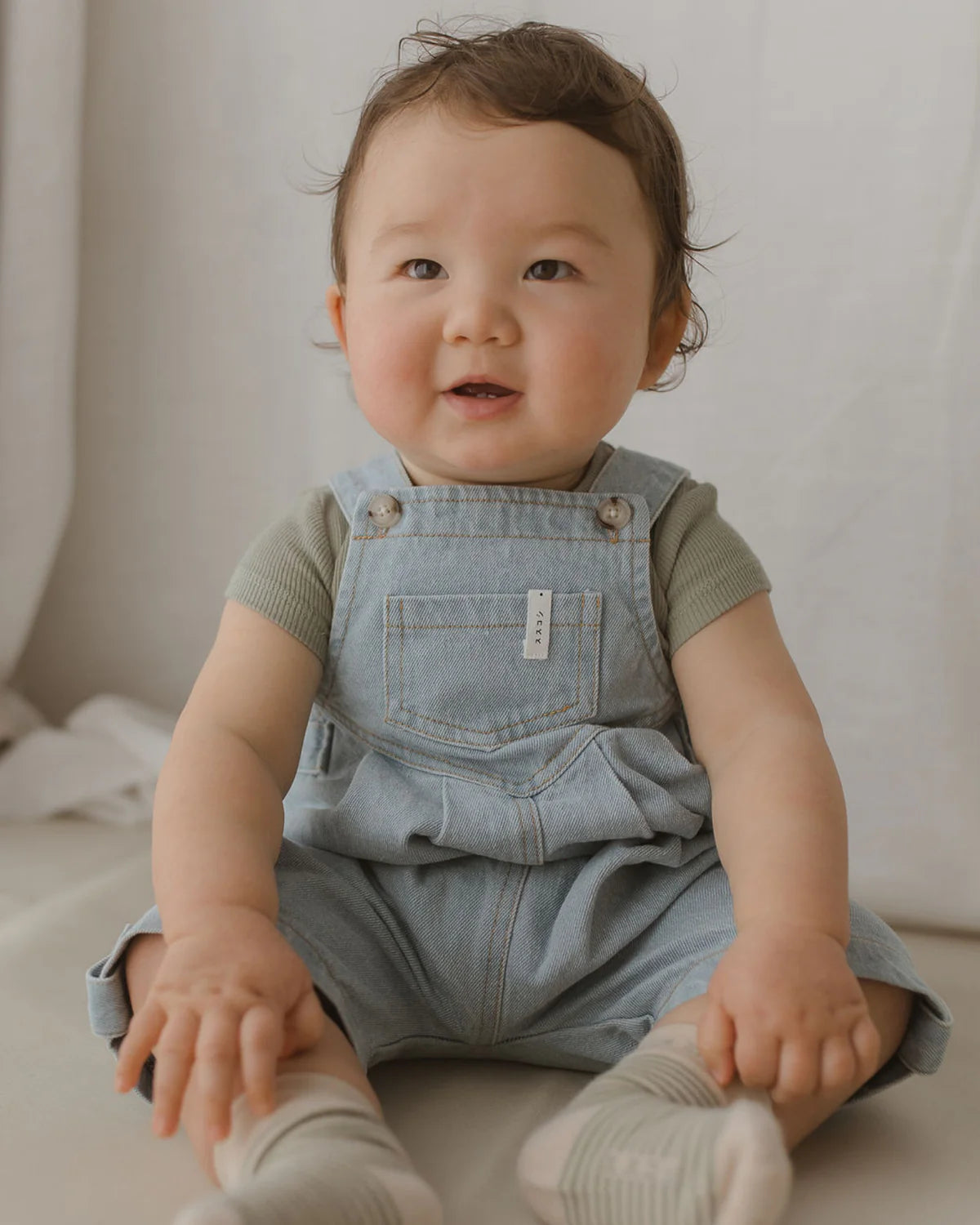 A baby with short, dark hair sits on a light surface, wearing SUSUKOSHI's Denim Short Overalls Baby Blue made from ethically-made organic cotton. The baby has a cheerful expression, with a slight smile and bright eyes, and is reaching forward with both hands.