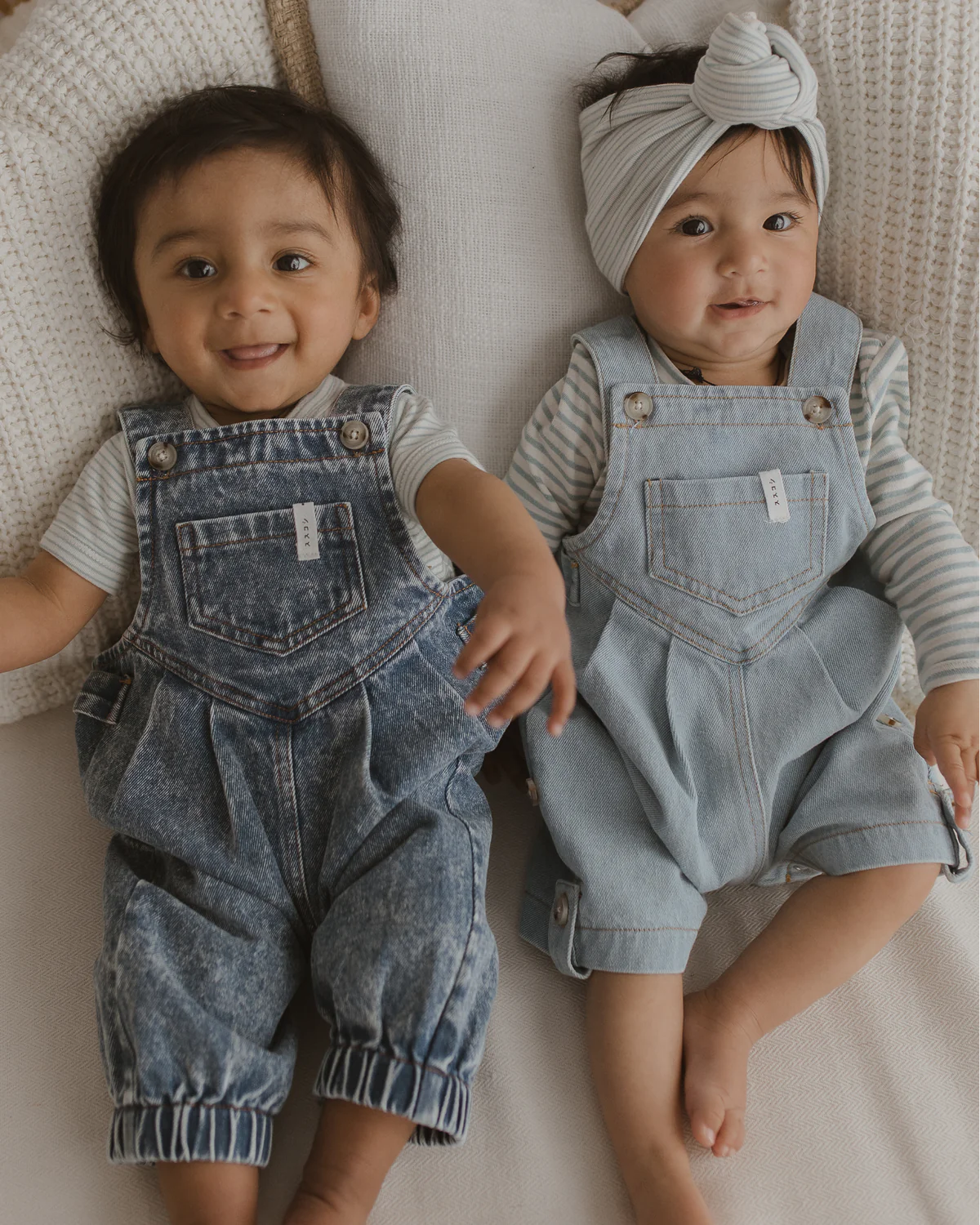 Two babies are sitting close together, wearing matching SUSUKOSHI Denim Short Overalls in Baby Blue, crafted from organic cotton, paired with light-colored striped shirts. The baby on the right is accessorized with a headband featuring a knot, while the baby on the left has no headband. Both babies are smiling and laying on a white, knitted blanket.