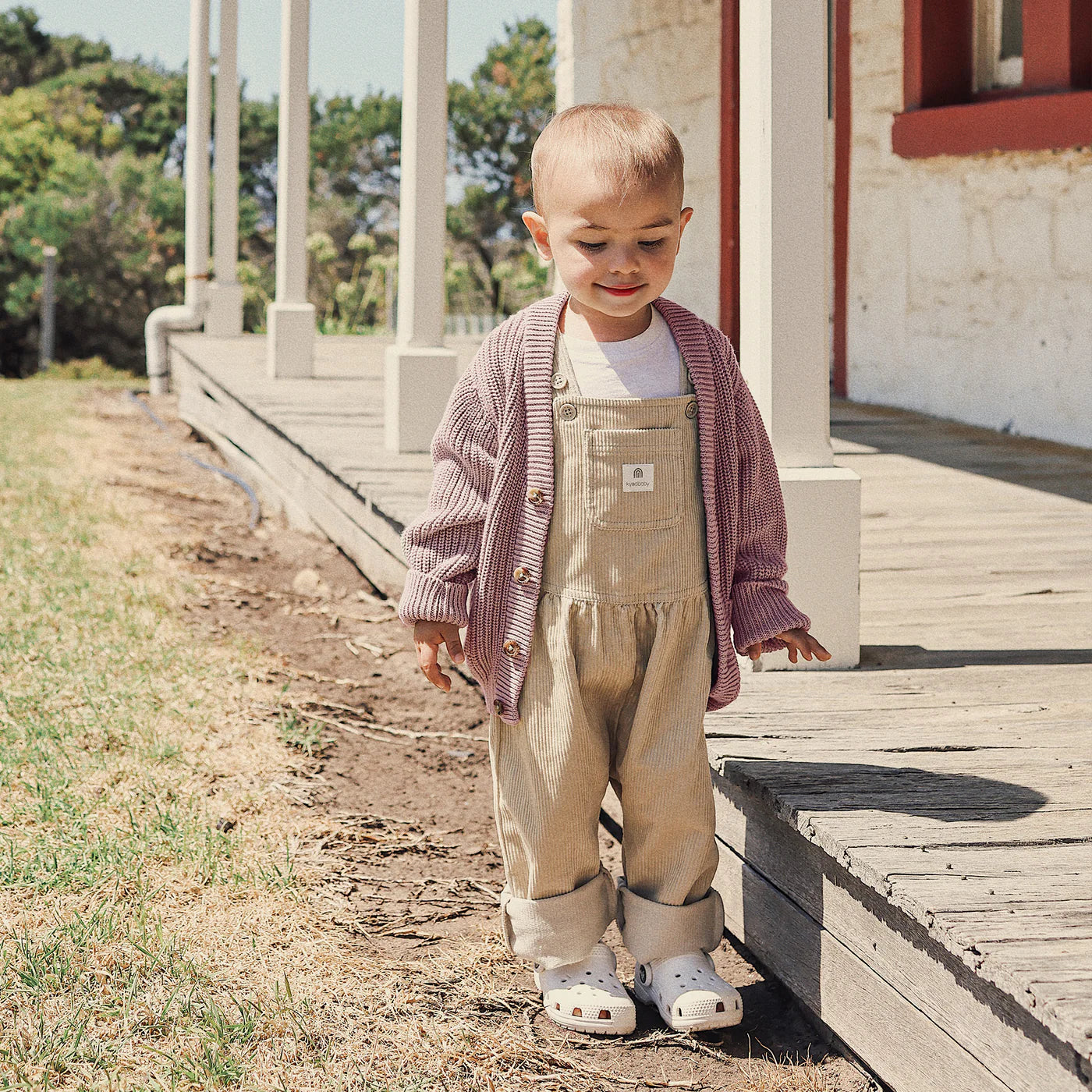 A young child stands by a wooden porch, wearing beige overalls, a white shirt, white shoes, and the KYND BABY Chunky Knit Cardigan in Dusty Lilac. The child smiles slightly while looking down; trees and a white building are in the background.