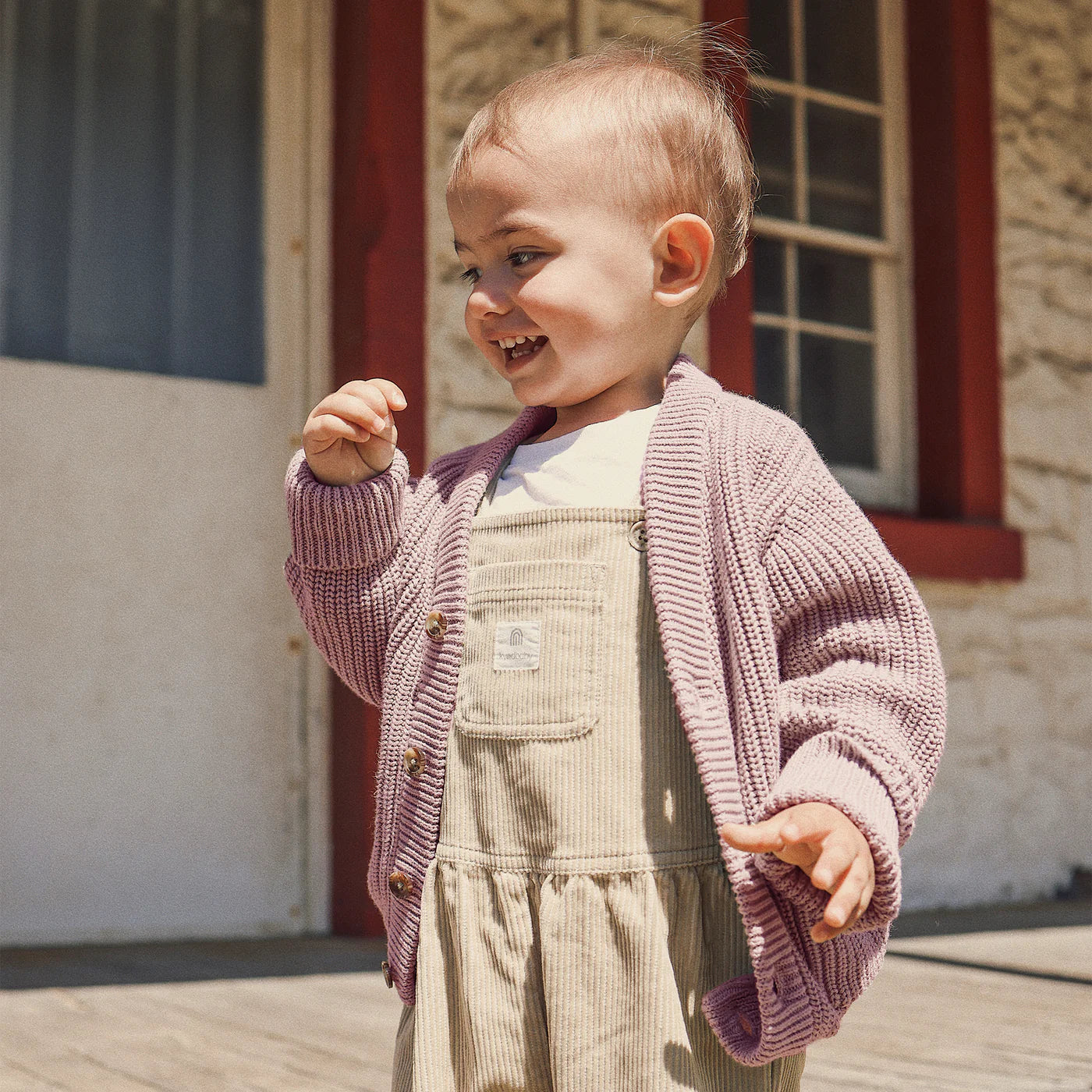 A smiling toddler wears the KYND BABY Chunky Knit Cardigan in Dusty Lilac, radiating comfort in front of a stone building with a door and window.