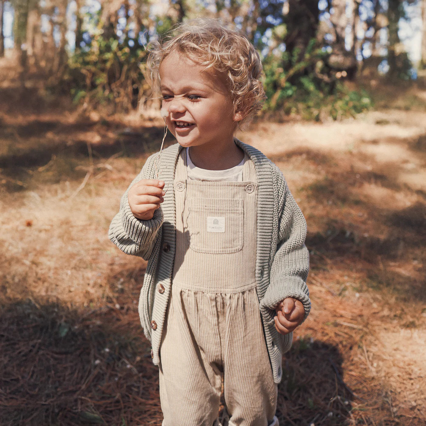 A smiling young child with curly blond hair stands outdoors in a sunlit forest, wearing the Kynd Baby - Chunky Knit Cardigan Sage from KYND BABY over beige overalls, ensuring comfort with its soft organic cotton. The child holds a small stick.