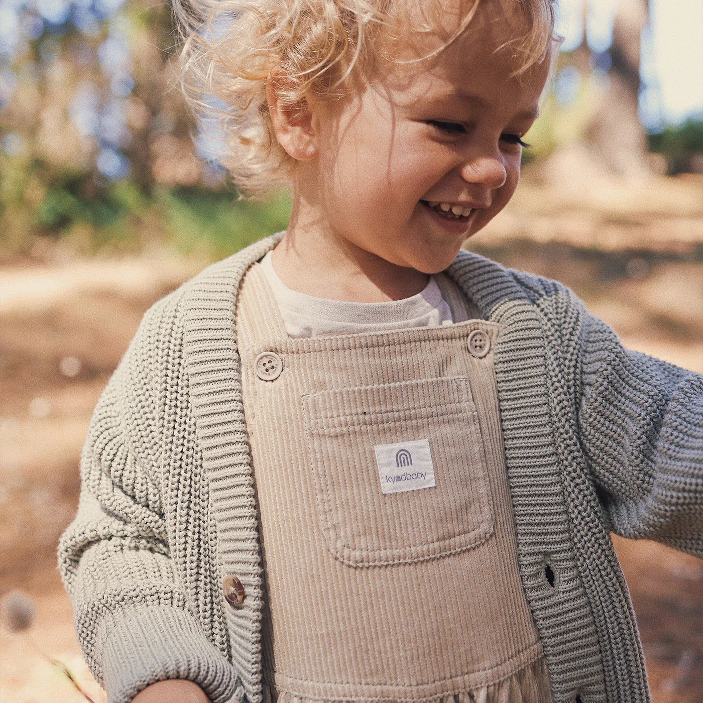 A smiling toddler wears the KYND BABY Chunky Knit Cardigan in Sage, crafted from soft organic cotton for ultimate comfort, layered over beige corduroy overalls while standing outdoors on a sunny day with trees in the background.