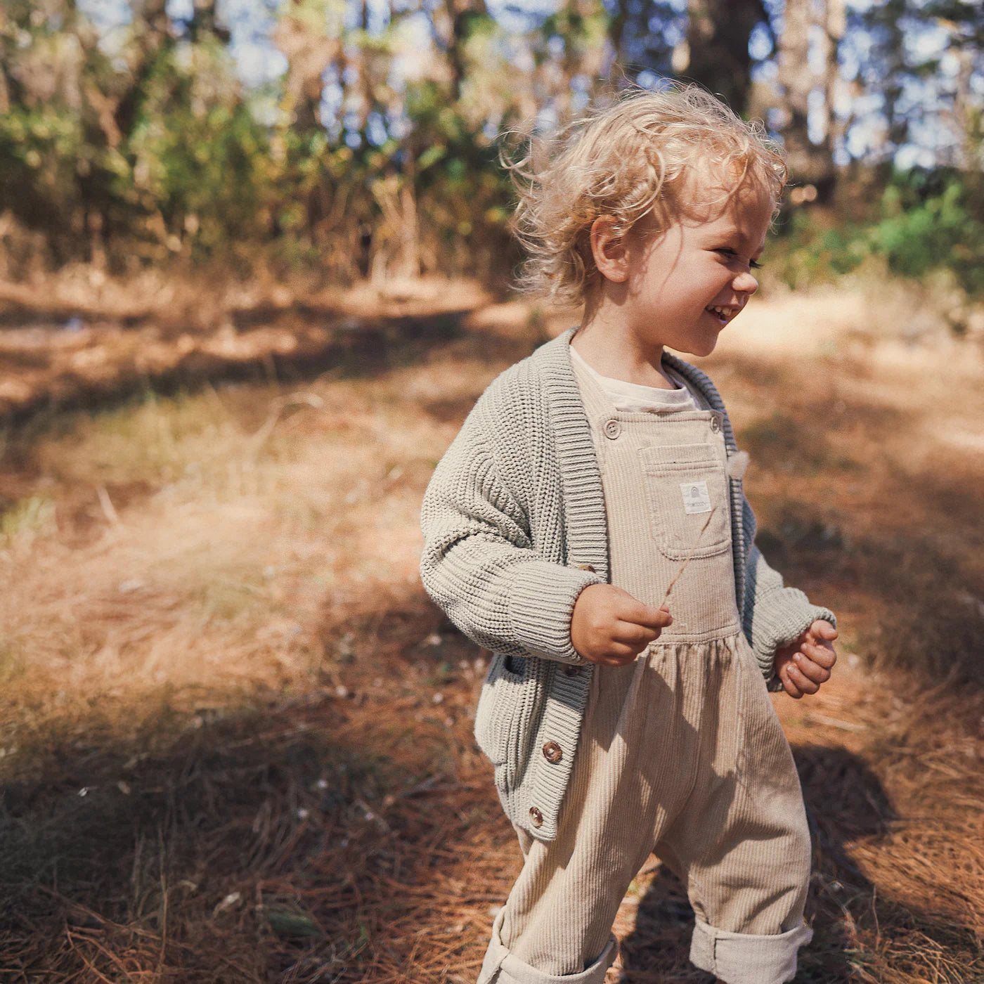 A young child with curly blonde hair, wearing the KYND BABY Chunky Knit Cardigan Sage, smiles while walking outdoors on a sunlit forest floor covered in pine needles.