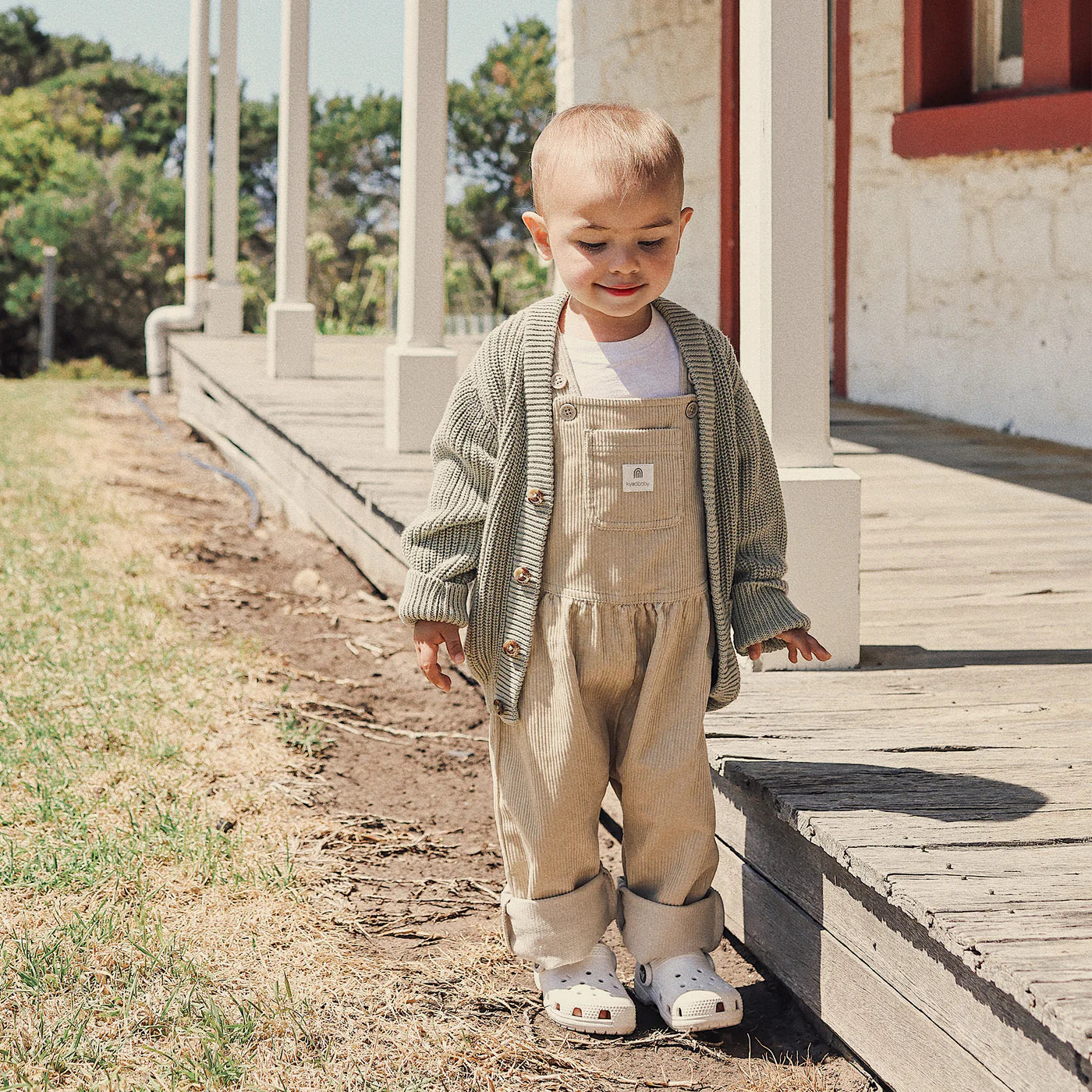 A young child stands on grass by a porch in the sunshine, wearing light overalls, a white shirt, and the KYND BABY Kynd Baby - Chunky Knit Cardigan Sage for cozy comfort. Trees and a building appear in the background.
