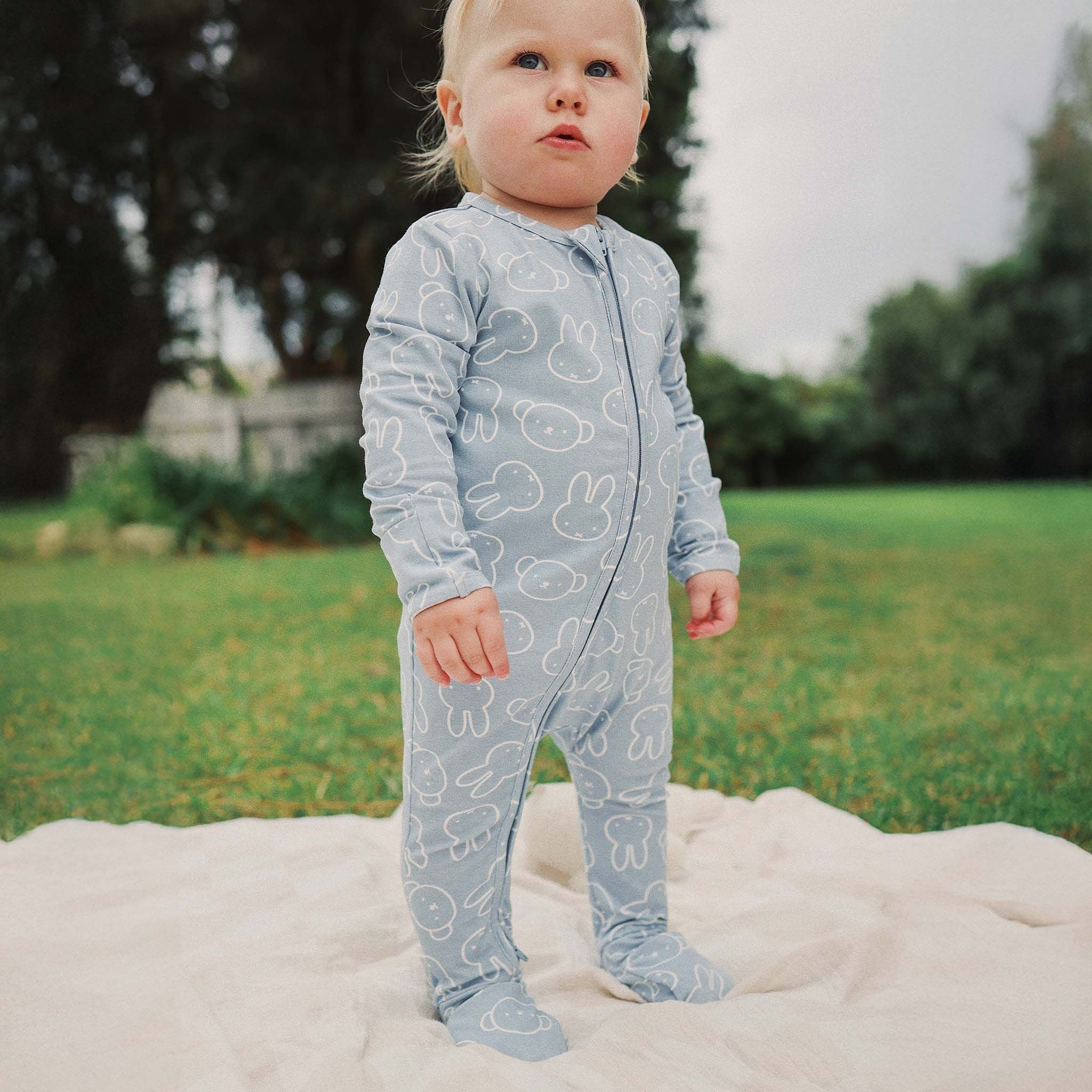 A toddler stands on a light-colored blanket outdoors, wearing the KYND BABY Kynd Baby - Miffy Day Or Night Onesie Moody Blue. Green grass and trees appear in the background.