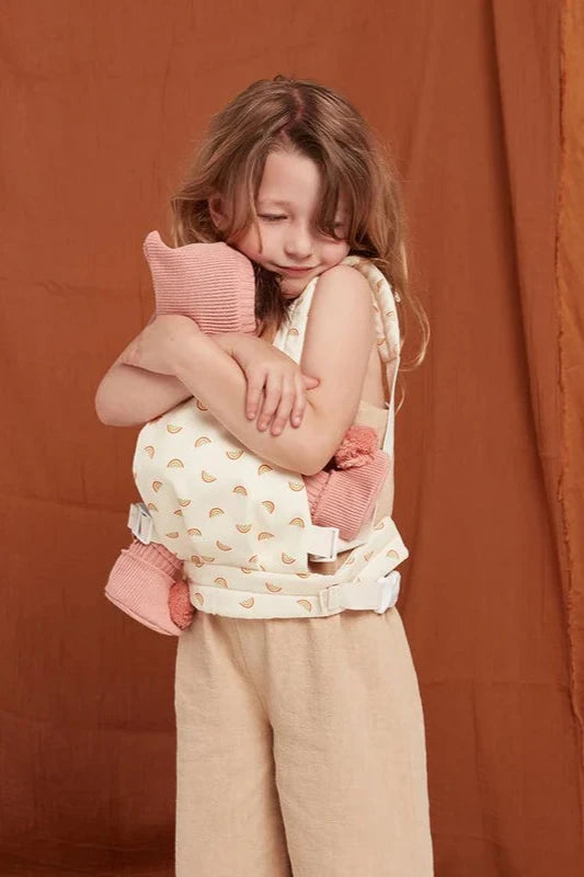 A young child with long hair is snuggling a stuffed animal while wearing a white Dinkum Dolls Rainbow Carrier with yellow patterns by OLLI ELLA. Standing against a brown background and dressed in light-colored pants and a sleeveless top, the child appears content and affectionate, embodying the joys of babywearing fun.