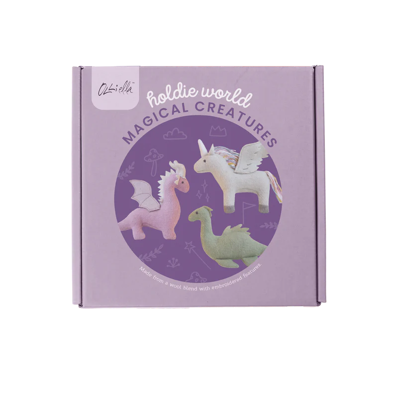 A lavender-colored box featuring the "Holdie Magical Creatures" label showcases illustrations of a pink dragon, Unicorn Dragon Nessy, and a green dragon. The box prominently displays the OLLI ELLA logo and highlights that the toys are crafted from a soft wool blend with embroidered features.