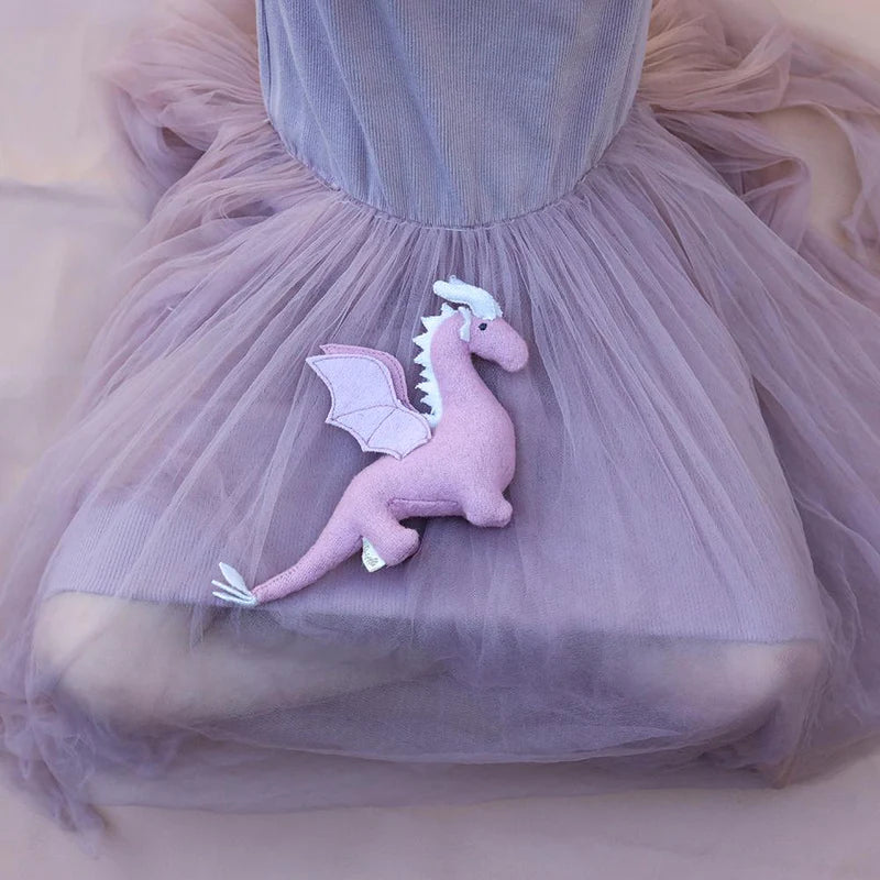 A person in a flowing, pastel pink tulle dress is sitting with their knees tucked under them. Resting on their lap is OLLI ELLA's Holdie Magical Creatures' Unicorn Dragon Nessy, a small, pink stuffed dragon toy with white details on its wings and tail.