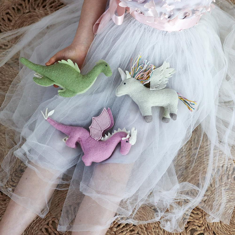 A child dressed in a white tutu-style skirt holds three OLLI ELLA's Holdie Magical Creatures: a green dinosaur, a beige unicorn with a colorful mane and tail, and a pink dragon with wings. The child is seated on a woven mat.