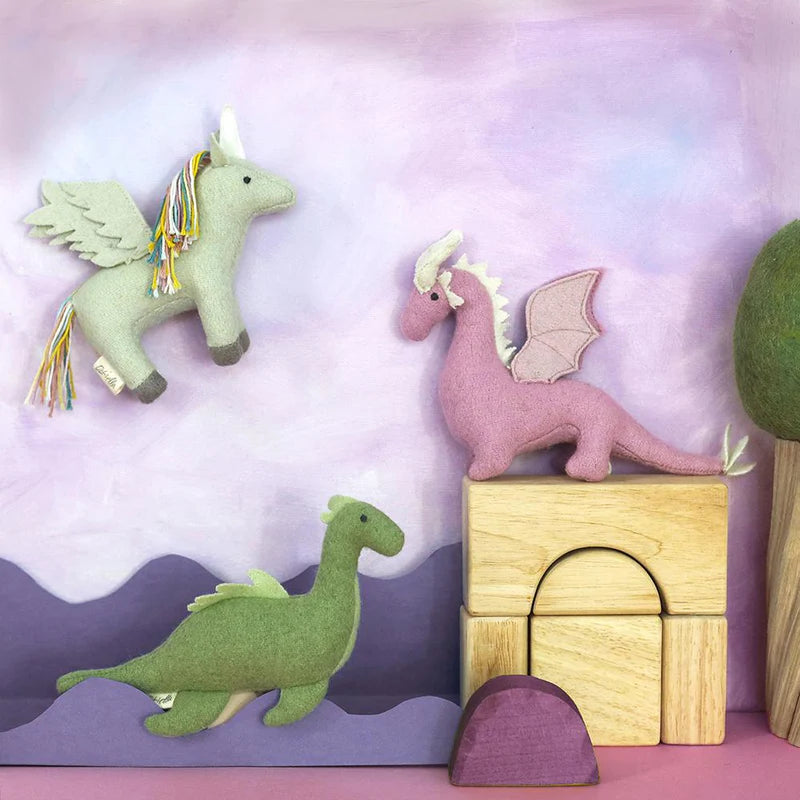 A playful scene featuring colorful felt toy dragons: a green one walking on a lilac surface, Holdie Magical Creatures' pink Unicorn Dragon Nessy with wings perched on wooden blocks, and a flying dragon with white wings and a rainbow mane above a purple hilly background. A felt tree stands on the right. All from OLLI ELLA's enchanting collection.