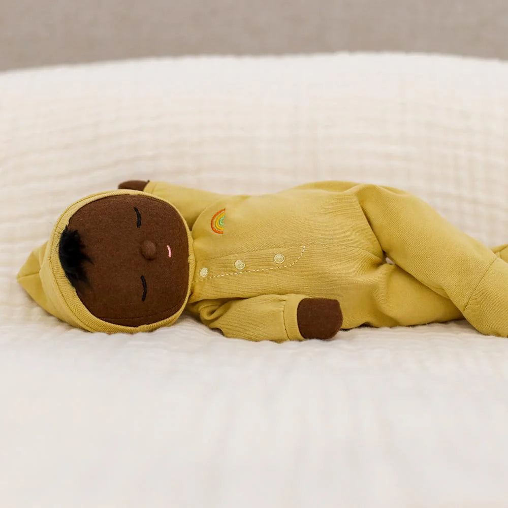 A Dozy Dinkum Mini soft doll from OLLI ELLA with smooth brown skin lies on a cream-colored, textured blanket. This baby’s first toy wears a yellow outfit with a hood and has a small rainbow embroidered on the chest. With its eyes closed and an embroidered face, it exudes a serene expression.