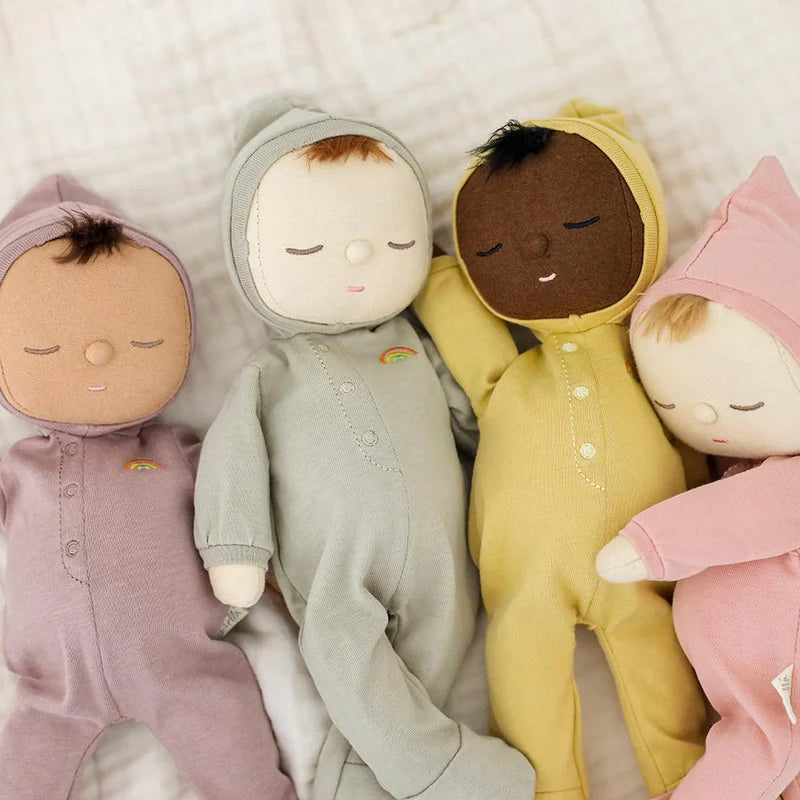 Four OLLI ELLA Dozy Dinkum Minis, dressed in pastel-colored onesies—purple, grey, yellow, and pink—are lying side by side on a soft surface. Each plush baby doll's first toy features an embroidered face with closed eyes and peaceful expressions, complete with small details like rainbows on their outfits.