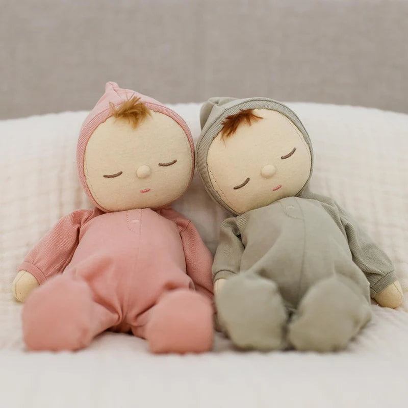 Two soft fabric dolls, named Dozy Dinkum Pickle from OLLI ELLA and dressed in cozy hooded onesies—one pink, one green—sit side by side on a quilted white surface. Each doll has closed eyes and a small tuft of brown hair peeking from under their hoods, perfect for cuddles and play as a baby's first toy.