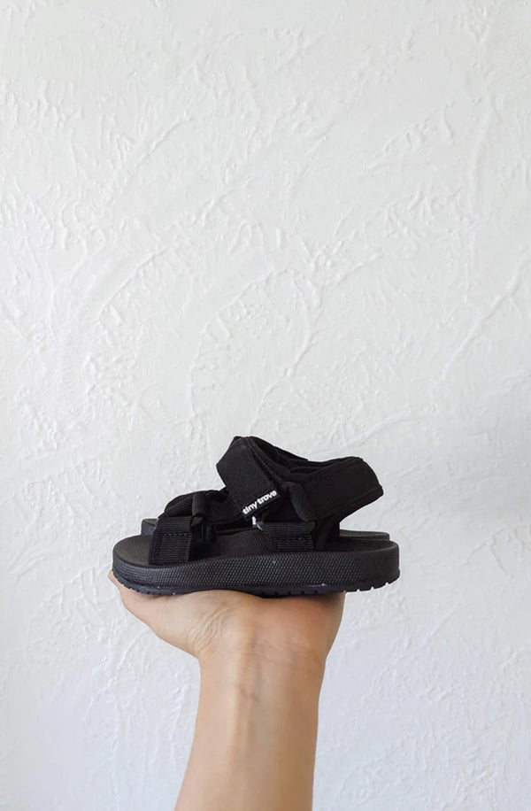 Hand holding the Olympi sandal in black against white textured wall