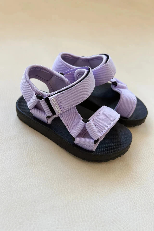 The Olympia Velcro Sandal Lilac by TINY TROVE showcases light purple straps paired with black soles, offering a flexible sole and adjustable Velcro for a comfortable fit. These sandals are elegantly displayed on a light beige surface.