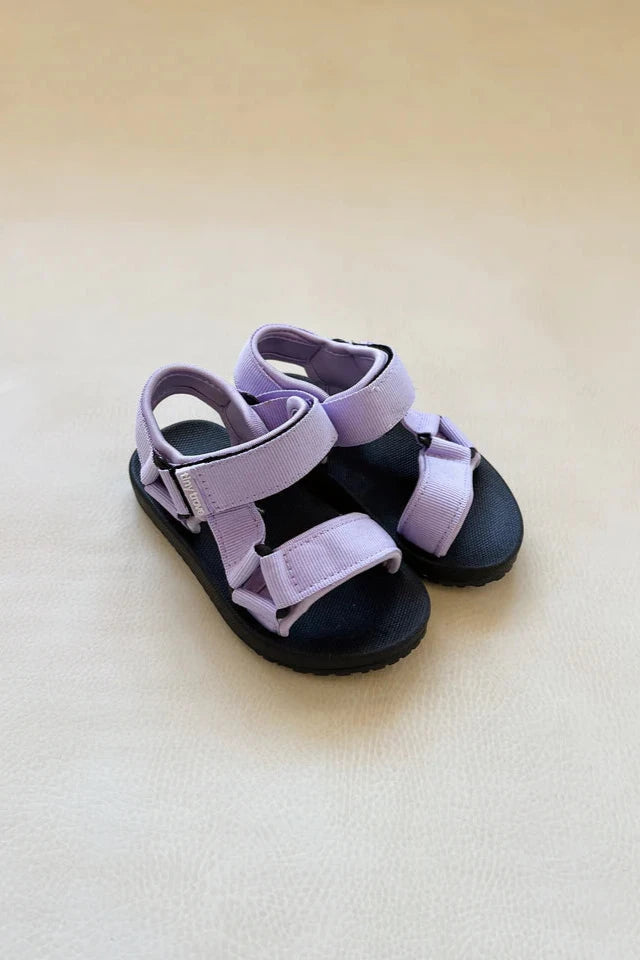 The Olympia Velcro Sandal Lilac by TINY TROVE is a pair of children's sandals with flexible black soles and light purple fabric straps. They feature a closed-toe design and adjustable Velcro fastening, with the Tiny Trove logo details adding extra charm, all showcased on a soft, off-white surface.