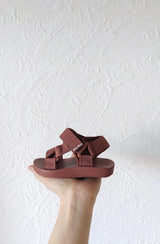 Hand holding the Olympia sandal in brick against white textured wall