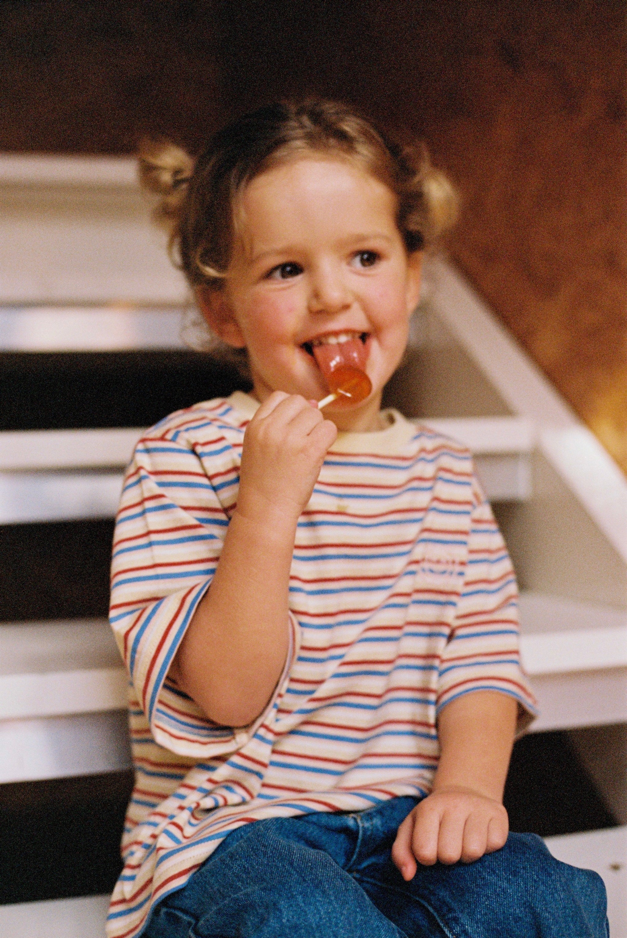 A young child with short, curly hair sits on white stairs, smiling and licking a lollipop. They are wearing an oversized Stripe Sloppy Tee from KOKOMO made from organic cotton and jeans, creating a cheerful and playful scene indoors.