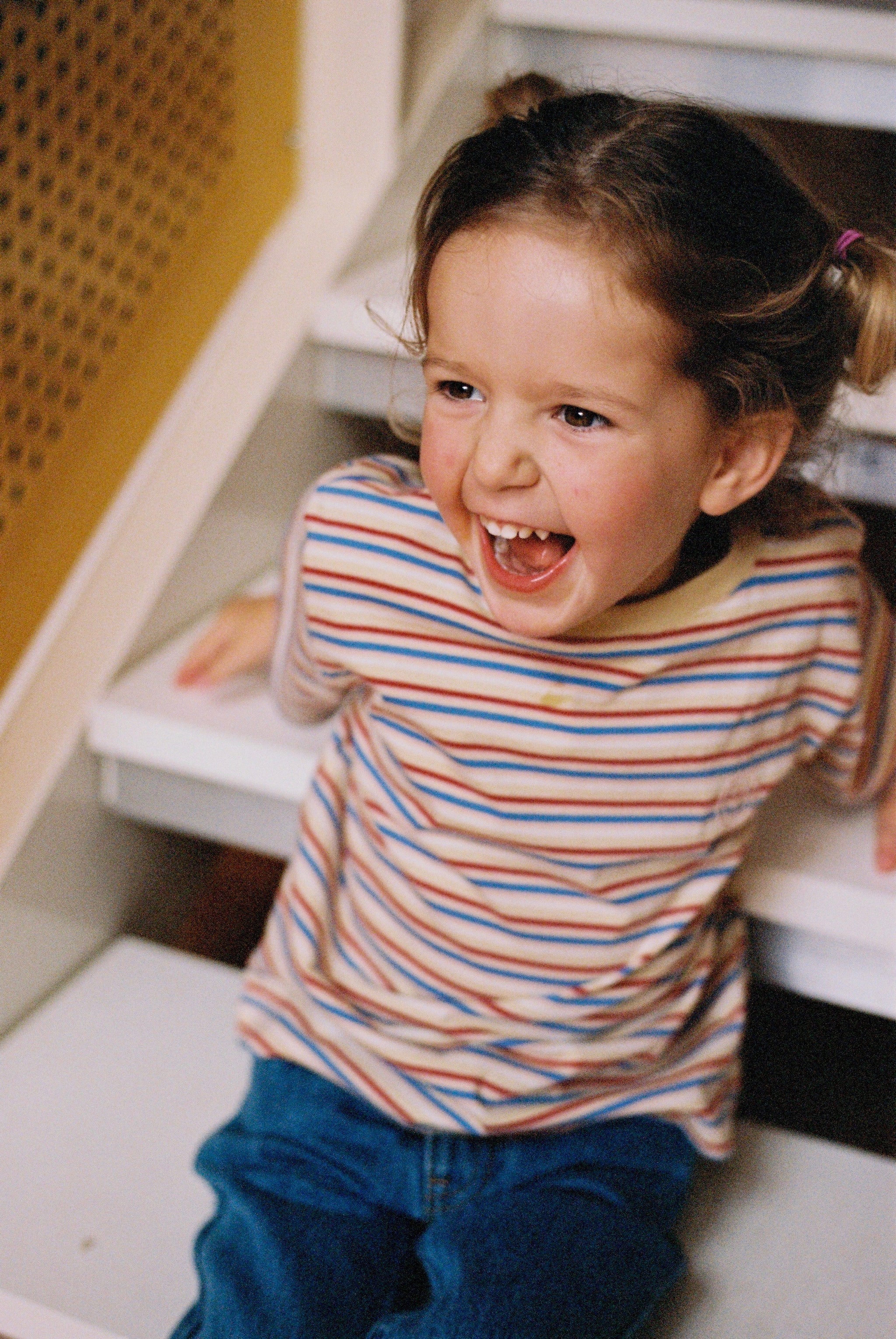 A young child with pigtails, wearing the oversized Stripe Sloppy Tee by KOKOMO made from organic cotton, sits on stairs. They are laughing joyfully and looking slightly to the side. The background features a section of a wall with a yellow mesh-like pattern.