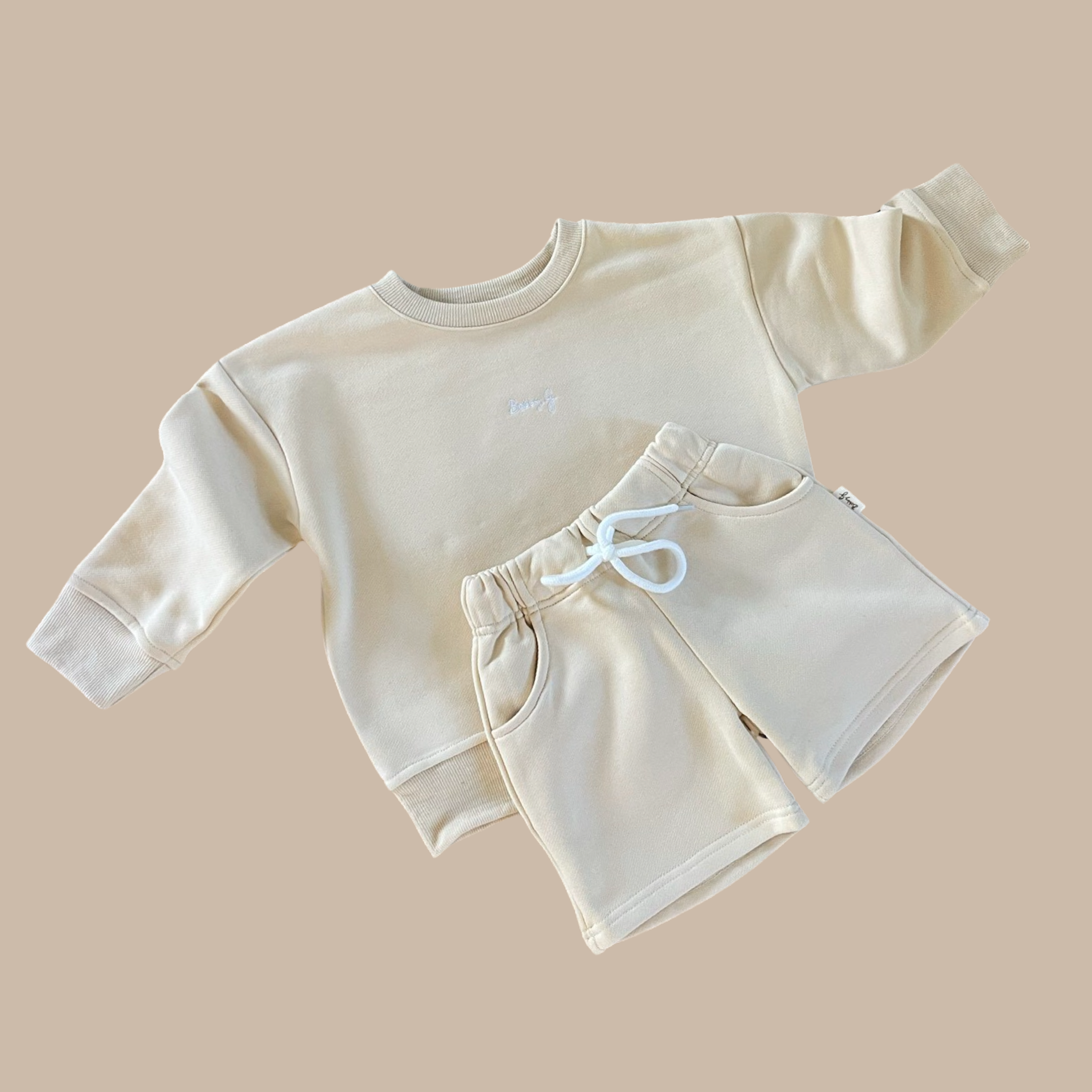 Against a neutral backdrop, the beige toddler outfit showcases BOBBY G BABY WEAR's "Frankie Shorts Popcorn" set. It includes a cozy French terry long-sleeve sweatshirt adorned with subtle "baby" embroidery, paired with matching jogger shorts featuring a white drawstring waist. Both pieces exude a soft and comfortable appearance.