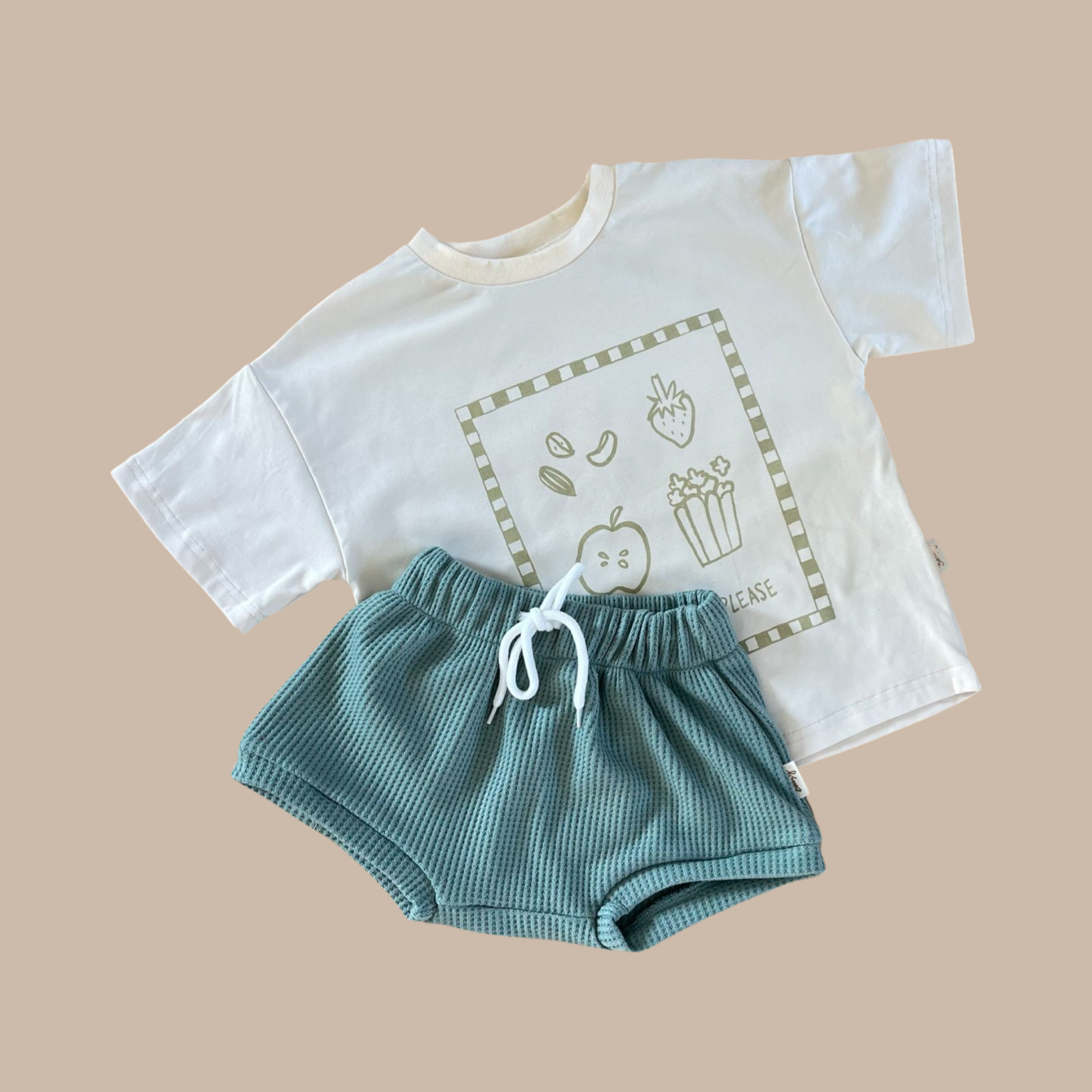 A 'More Snacks Please' Banana Tee from BOBBY G BABY WEAR, featuring fruit illustrations and made of organic cotton, is laid flat next to a pair of textured teal children's shorts on a beige background.