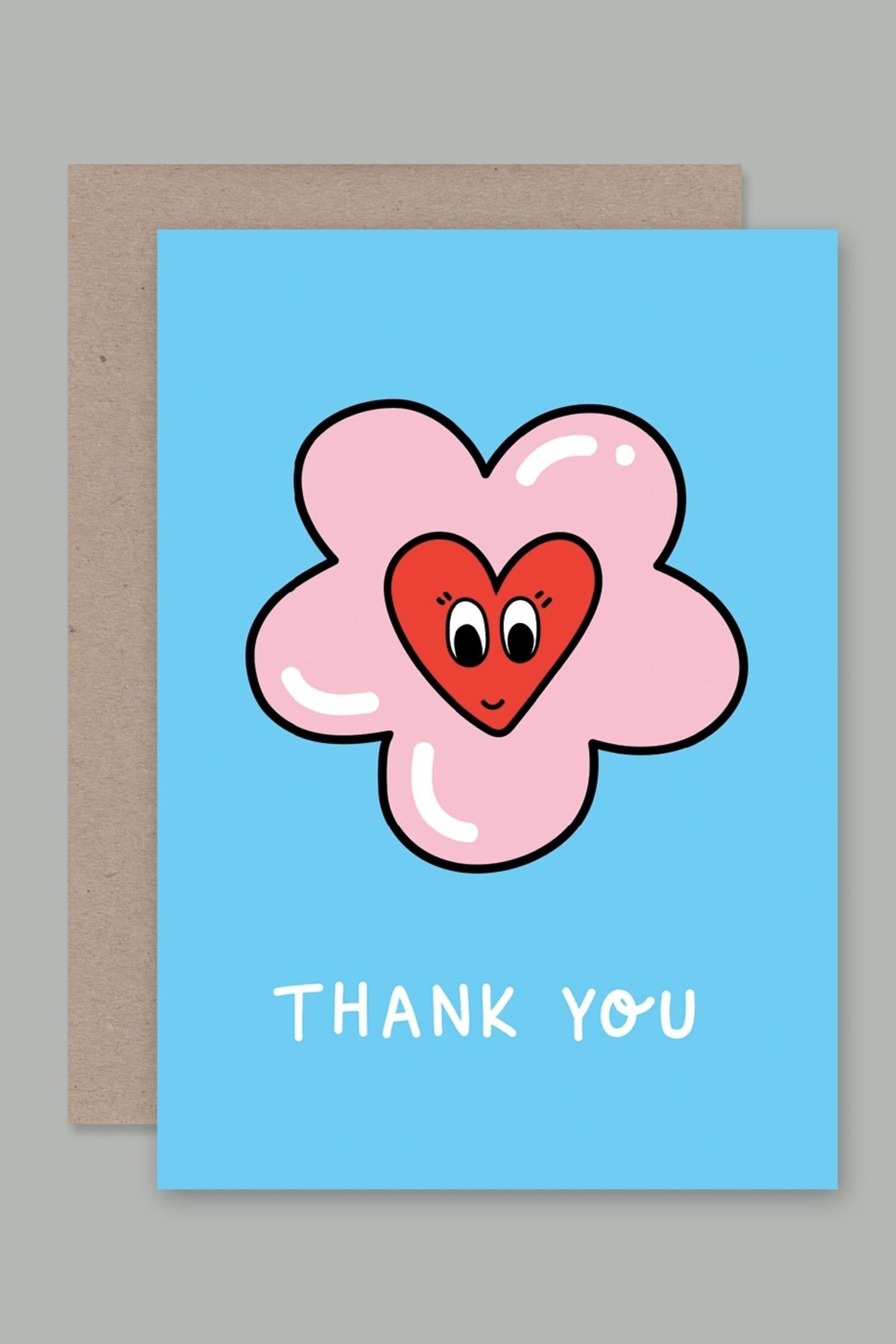 AHD PAPER CO's "Thank You" Greeting Card, crafted in Australia, displays a charming illustration of a pink flower with a red heart-shaped center that has eyes and a smile. The full-colour front boasts a light blue background with "THANK YOU" written in white at the bottom. The card is presented alongside a beige envelope.