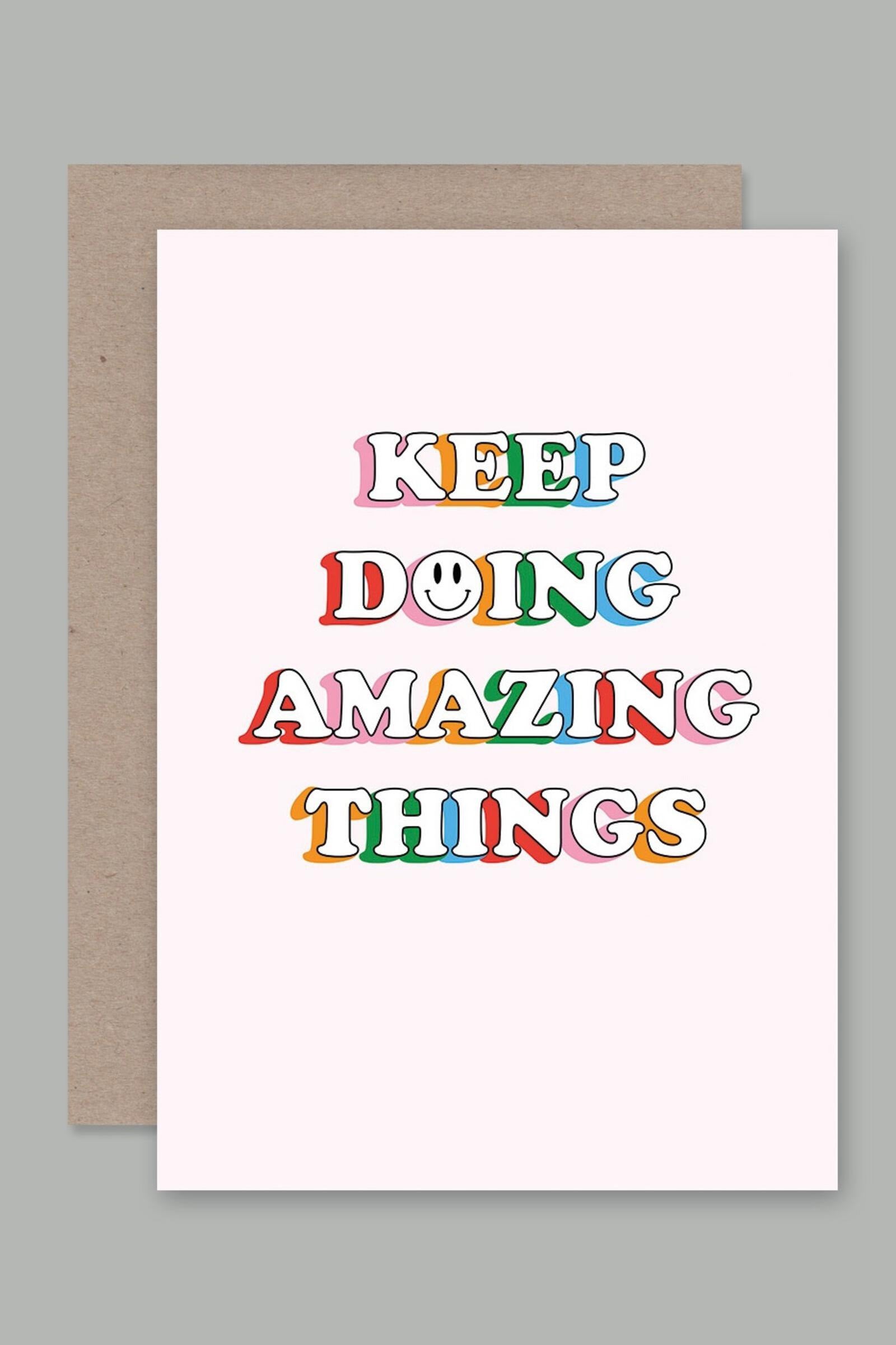 The **Greeting Card "Keep Doing Amazing Things"** by AHD PAPER CO, made in Australia, showcases colorful, whimsical text that states "KEEP DOING AMAZING THINGS" in uppercase letters. The letter "O" in "DOING" features a smiley face. Crafted from recycled paper, the card is elegantly presented on a brown envelope against a light gray background.