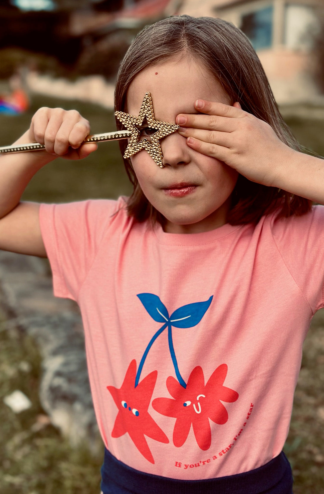 A young girl wearing the PLAYetc
You're a Star Tee holding a wand with a star on the end of it over her eye.