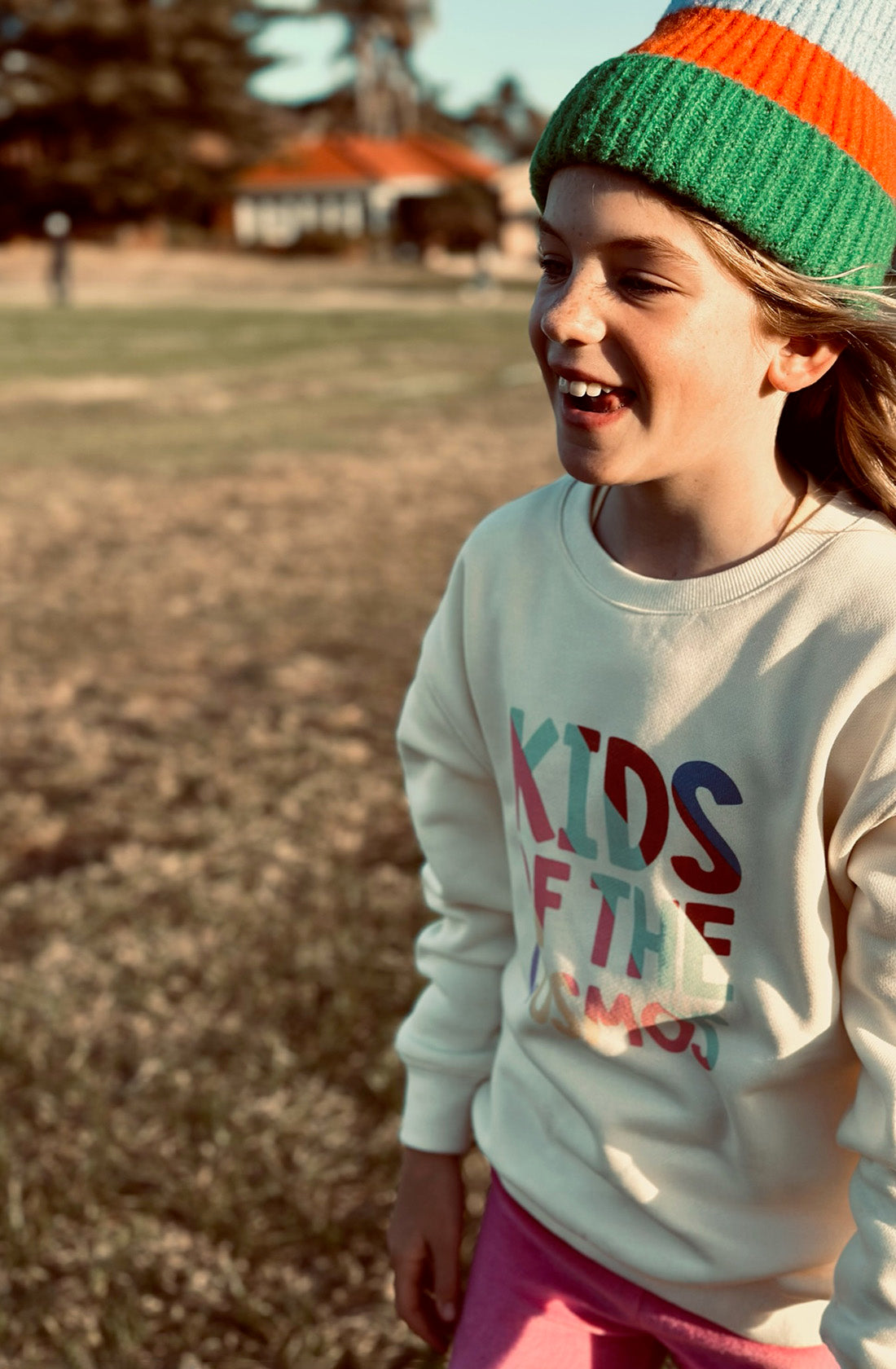 A young girl wearing the PLAYetc
Kids Of The Cosmos Sweater with a green, orange and blue beanie with pink pants running outside.