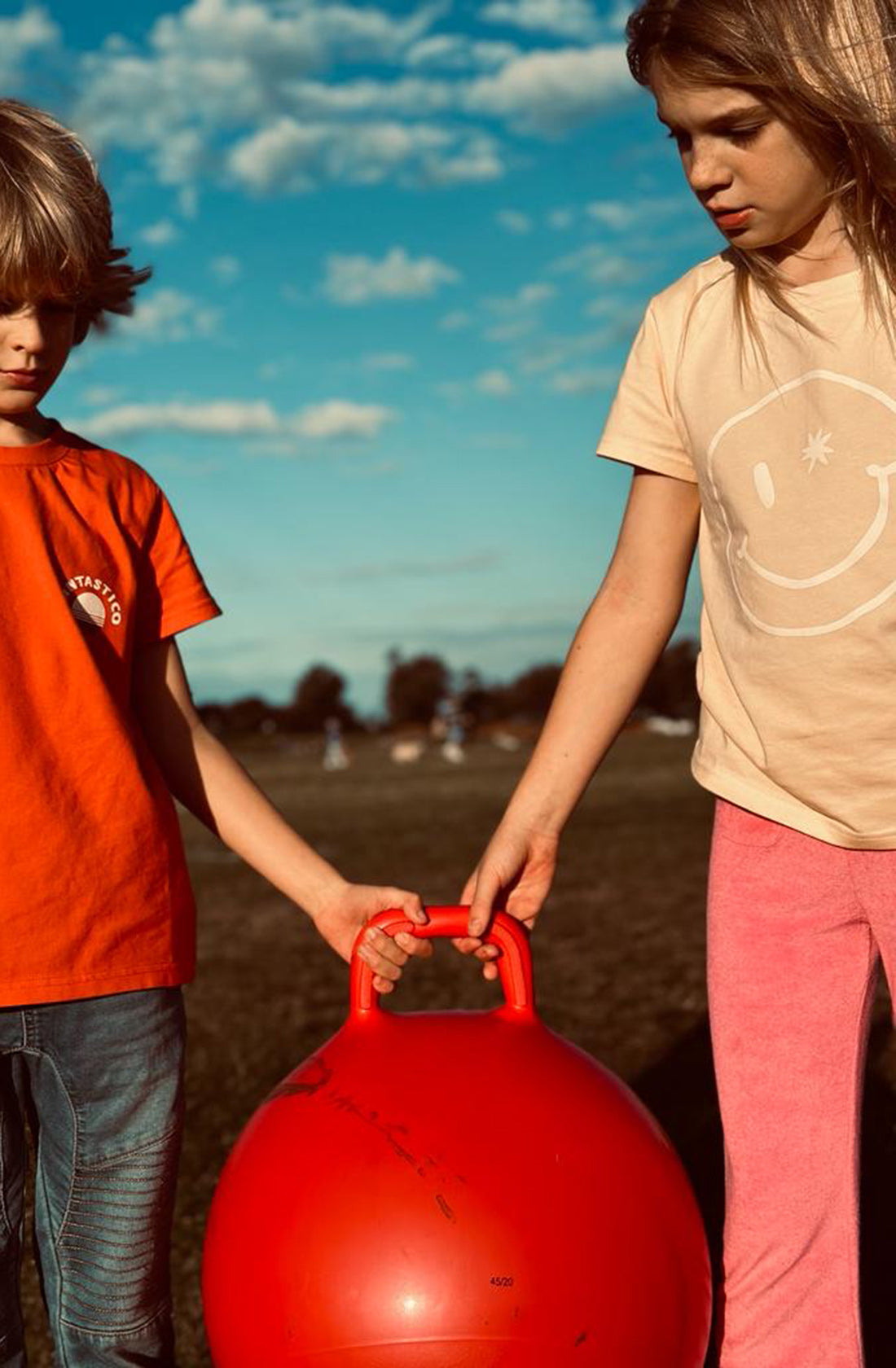 A young girl wearing the PLAYetc
Smiley Tee with pink pants holding a bouncey ball with another boy.