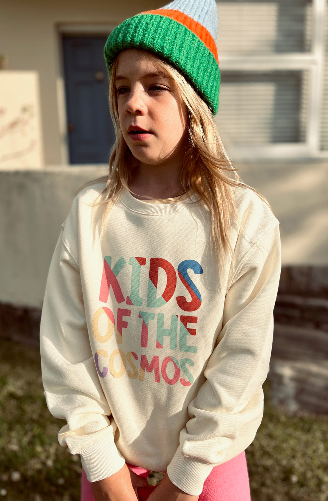 A young girl wearing the PLAYetc Kids Of The Cosmos Sweater with a green, orange and blue beanie with pink pants.