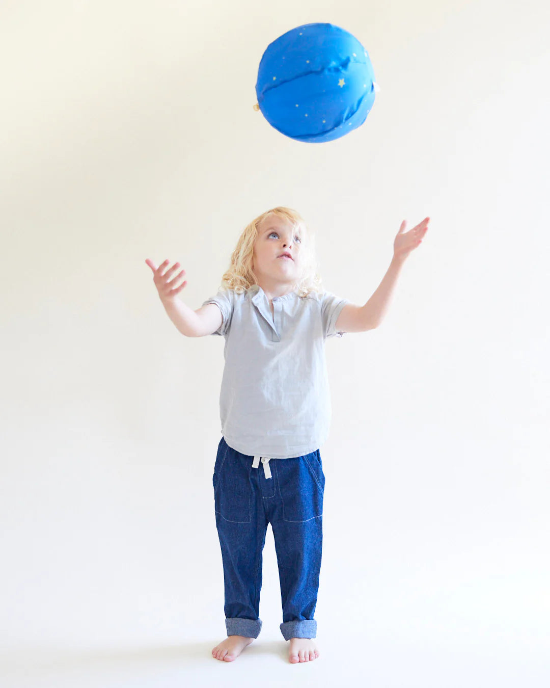 A child with blond hair and a light gray shirt, standing barefoot on a white background, is playfully tossing the Starry Night Balloon Ball by SARAH'S SILKS. Decorated with small stars, this reusable blue balloon ball captures the child's focused attention.