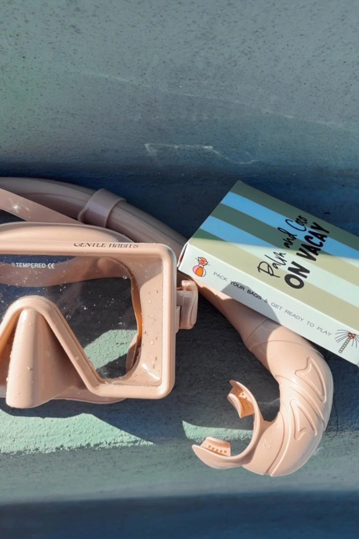 A beige snorkel and matching mask rest next to a striped "Lulu Pawlik - Palm & Coco On Vacay Kids Playing Cards" box by LULU PAWLIK, featuring hand-drawn art, on a textured gray surface in bright sunlight.