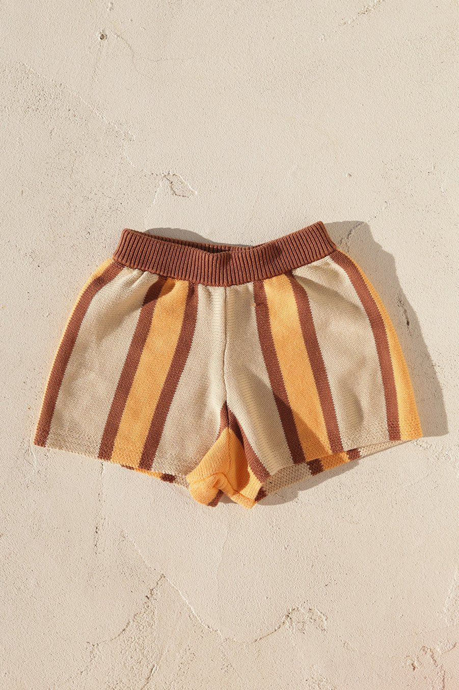 The Stripe Knit Shorts by SMALL SWIM CLUB boast a comfy loose fit and feature striking vertical stripes in brown, beige, and yellow, laid flat on a textured beige surface.