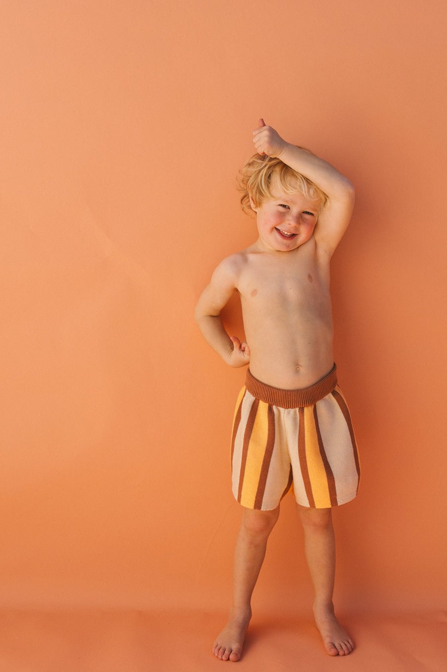 A smiling child with blond hair stands against an orange background, wearing Stripe Knit Shorts from SMALL SWIM CLUB. The comfy, loose-fit unisex cotton knit shortie features shorts with eye-catching stripes in shades of brown, yellow, and white. One arm is raised above their head, highlighting the vibrant color combination.