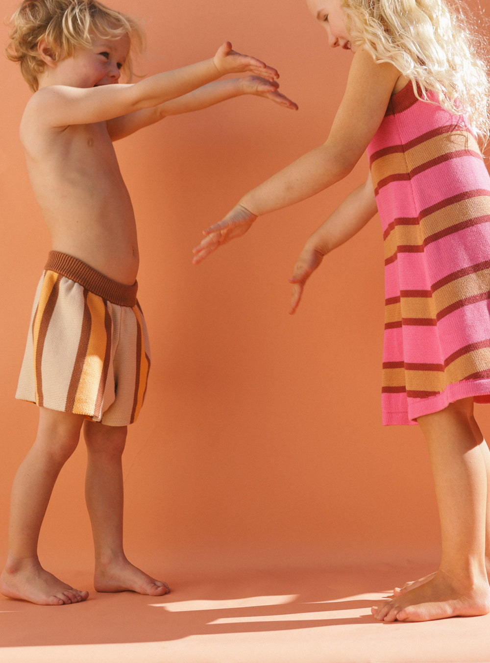 Two children playfully reach towards each other against an eye-catching peach-colored background. The child on the left wears the Stripe Knit Shorts by SMALL SWIM CLUB, while the child on the right sports a pink and brown striped dress. Both appear delighted and engaged in playful interaction.