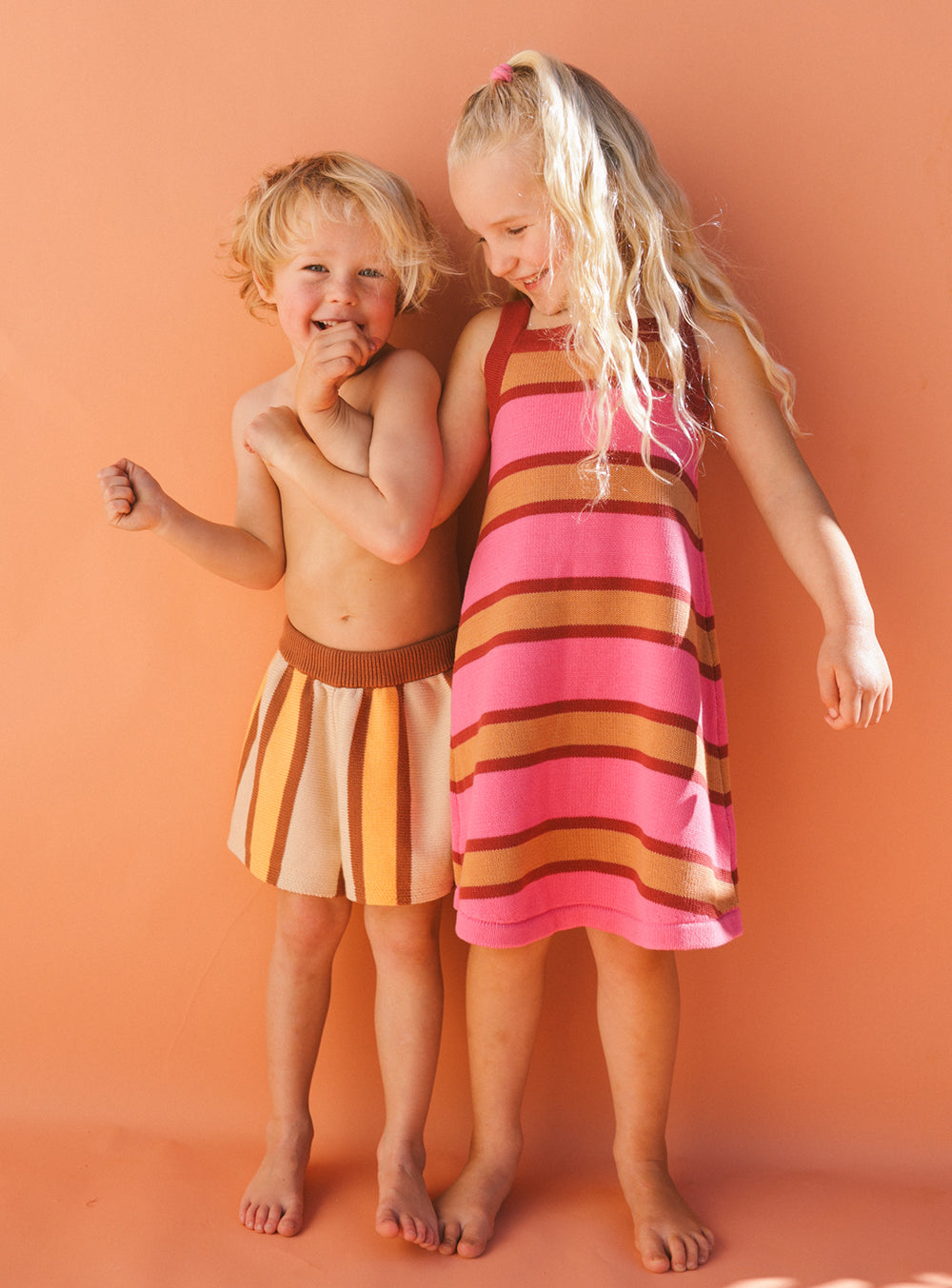 Two smiling children stand against an orange background. The child on the left wears a striped skirt in shades of brown, and the one on the right shows off a Stripe Knit Dress from SMALL SWIM CLUB in pink and brown. Both are barefoot and appear happy.