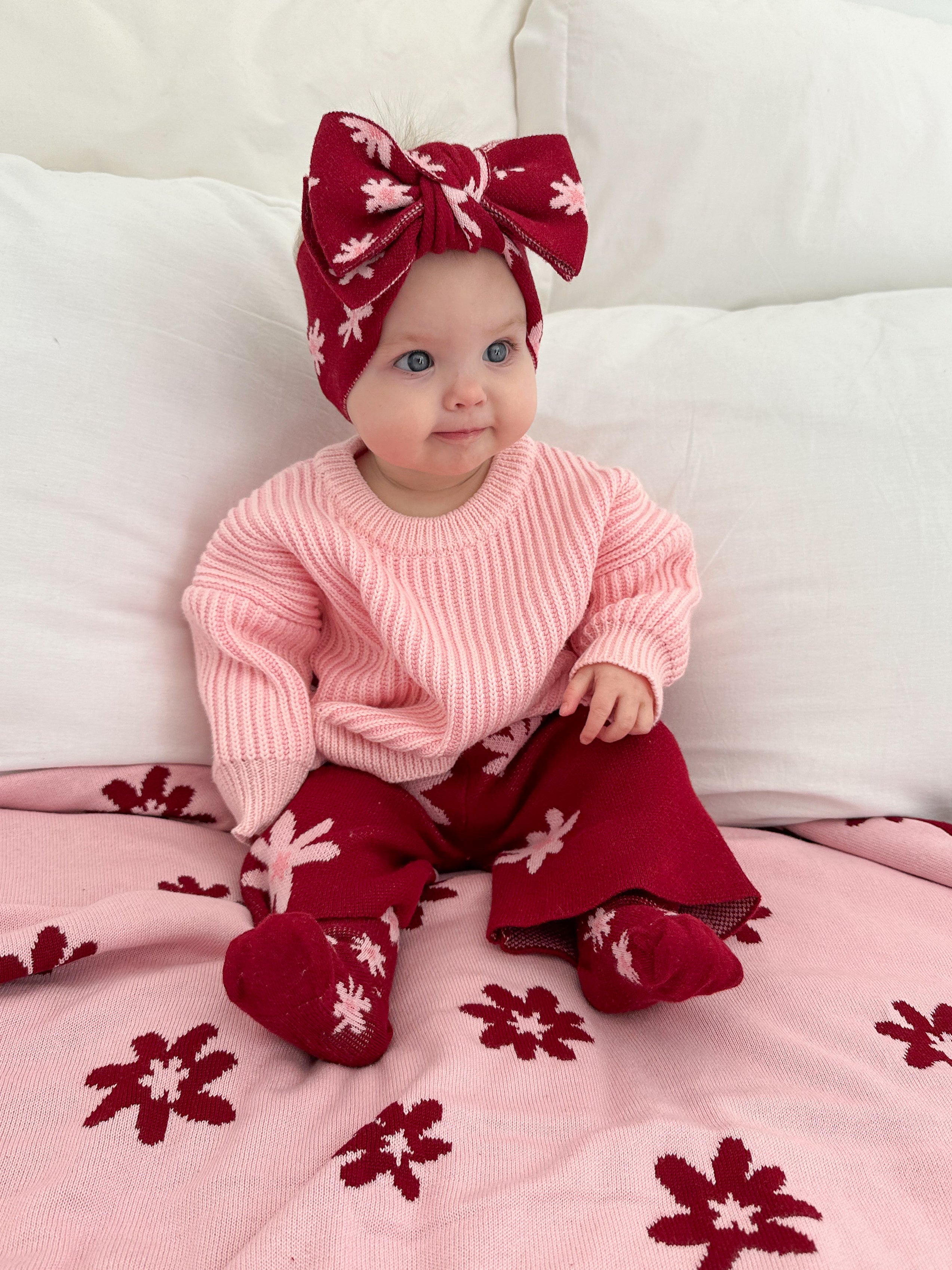 A baby wearing Ziggy Lou's Socks Lola (Plum) by ZIGGY LOU, a pink sweater, maroon pants, and a large floral headband sits on a bed with a pink floral blanket, smiling and looking slightly to the side.