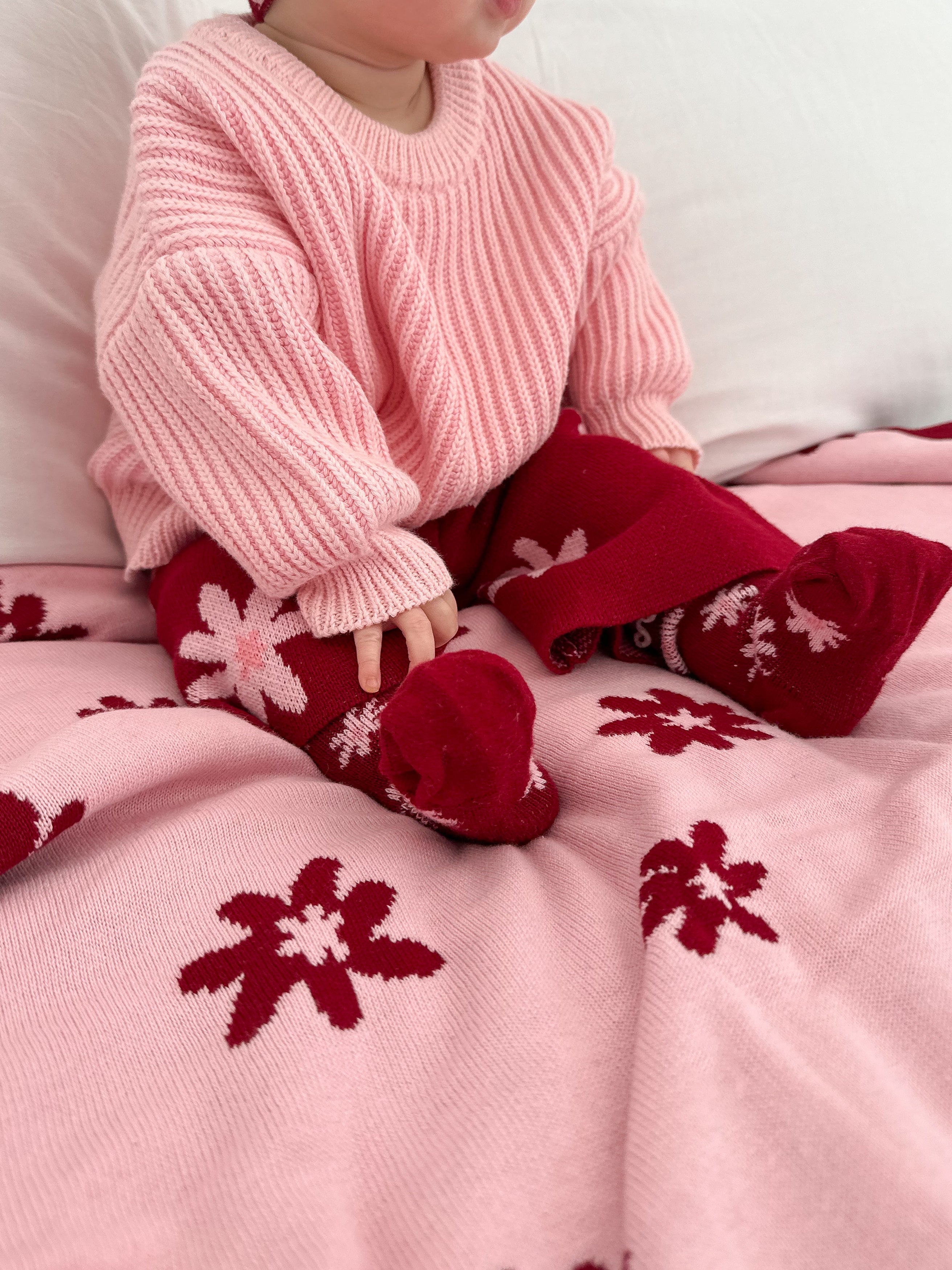 A baby sits on a pink blanket with red and white flower patterns, wearing ZIGGY LOU's Ziggy Lou - Socks Lola (Plum), a light pink knit sweater, and red tights with white flowers. The baby's face is partially out of frame.