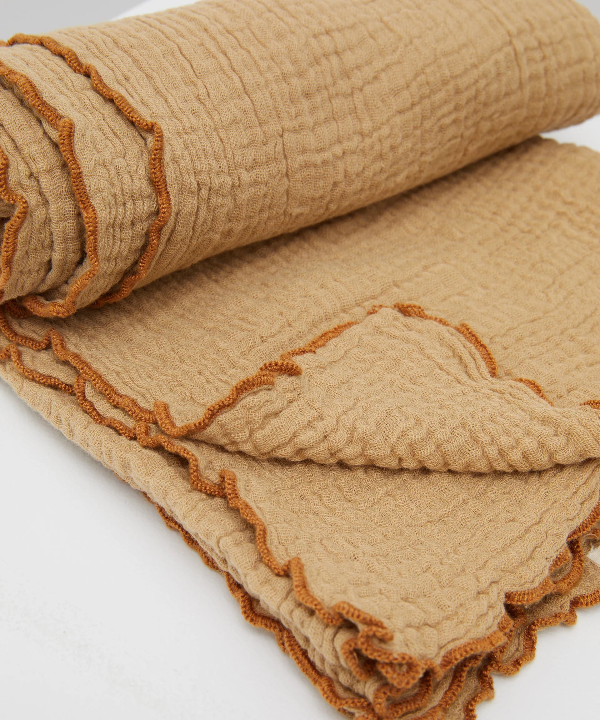 The Swaddle Cacao by PHILÉ, from the Cotton Classics Collection, is an extra-large muslin wrap in textured beige with a darker wavy edge pattern. Made of Oeko-Tex Certified Cotton, it looks soft and cozy folded on a plain white surface.