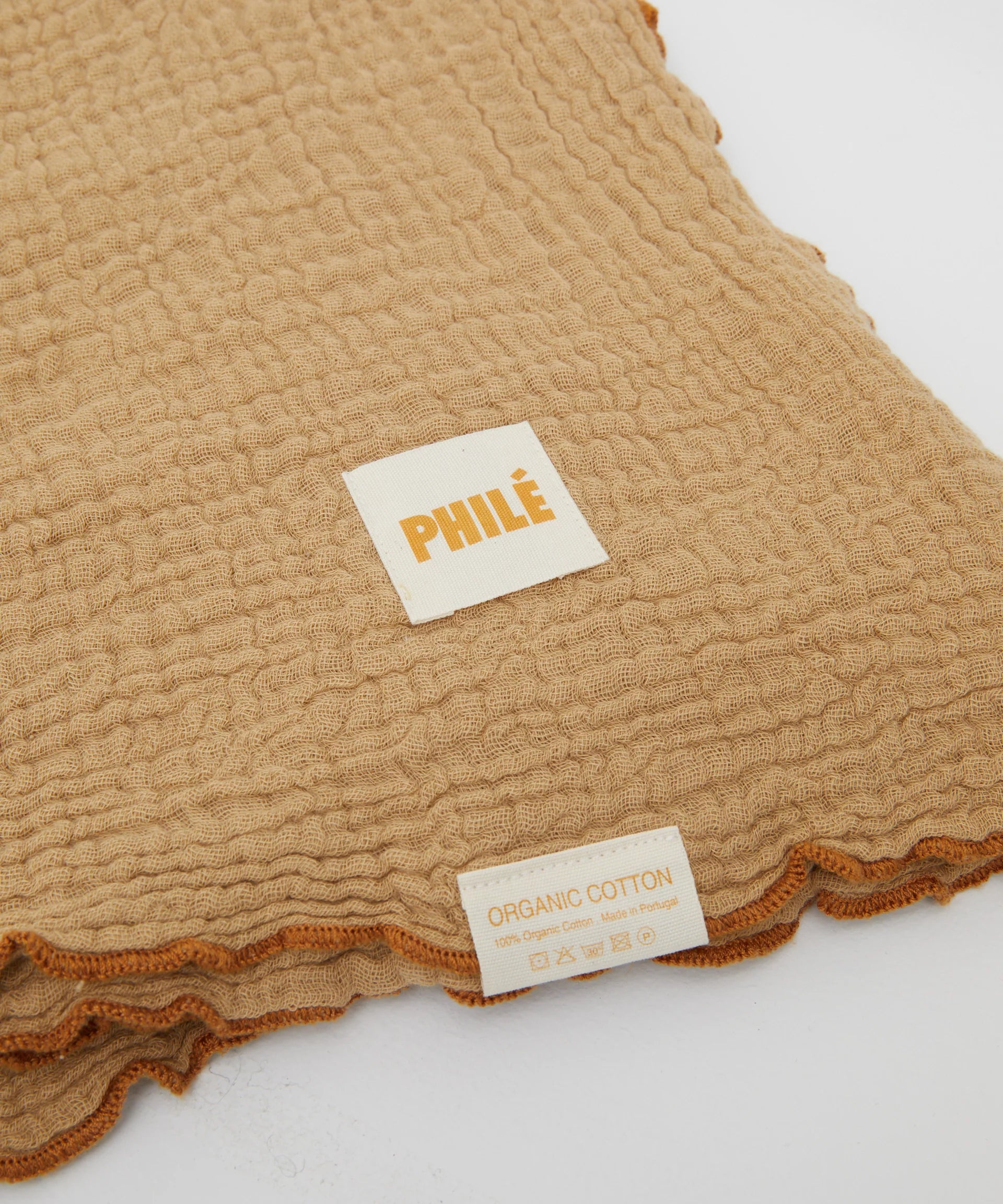 The Swaddle Cacao by PHILÉ is a textured beige fabric featuring an "ORGANIC COTTON" label and care symbols. It's made from Oeko-Tex Certified Cotton, part of the Cotton Classics Collection, with elegant orange-trimmed edges.