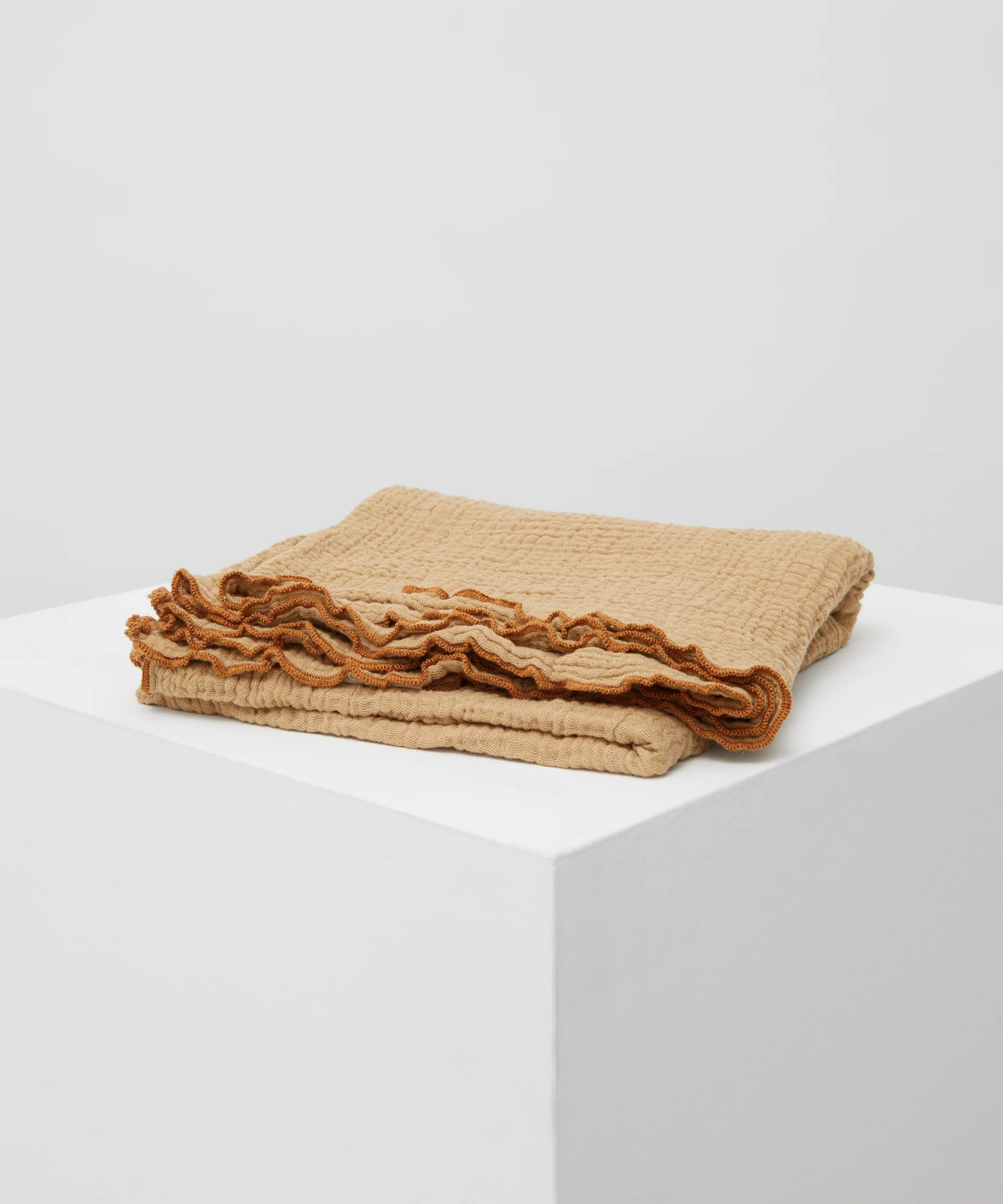 The PHILÉ Swaddle Cacao, a folded beige blanket with orange trim from the Cotton Classics Collection, sits on a white square pedestal against a light grey backdrop, showcasing its textured pattern and Oeko-Tex Certified Cotton for a cozy and stylish look.