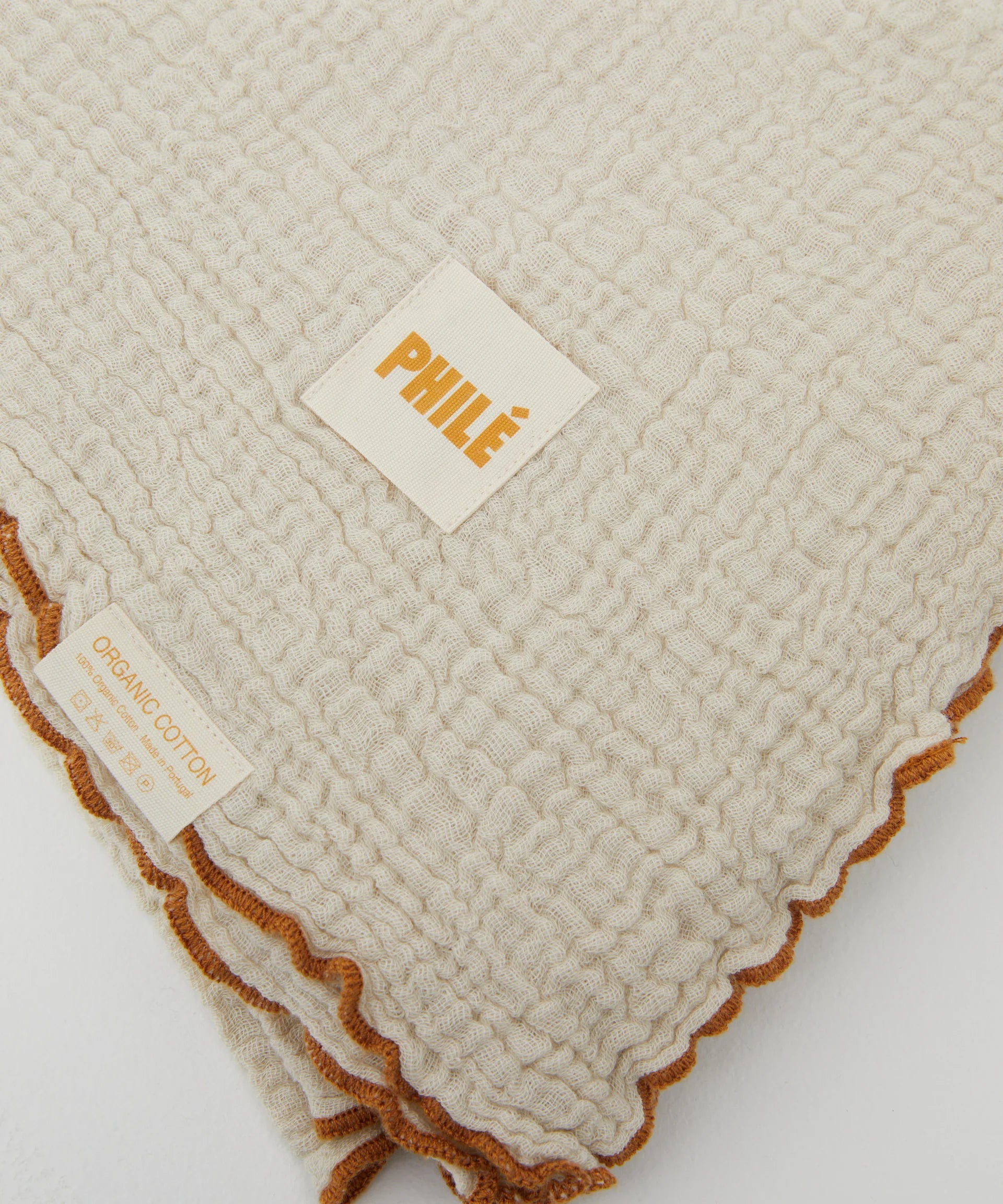 The Swaddle Crema from PHILÉ is a textured beige cotton blanket with an orange-trimmed edge. It features a small fabric tag reading "PHILÉ" in orange and uses Oeko-Tex Certified Cotton, ensuring organic materials.