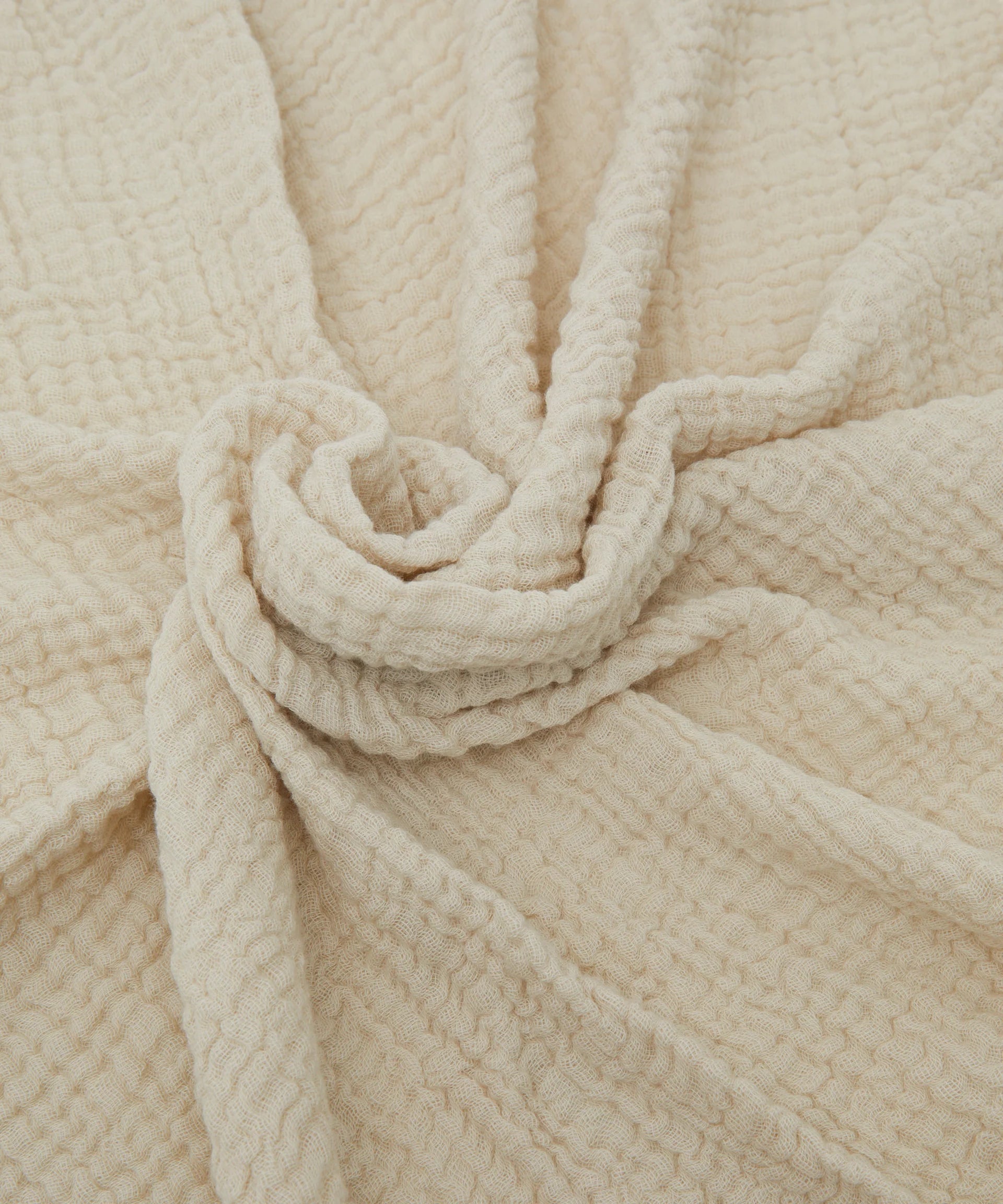 Close-up of PHILÉ's Swaddle Crema, a soft, beige textured fabric with a wrinkled pattern and central knot. Part of the Cotton Classics Collection, this plush material suggests warmth and comfort, ideal for an extra-large muslin wrap.