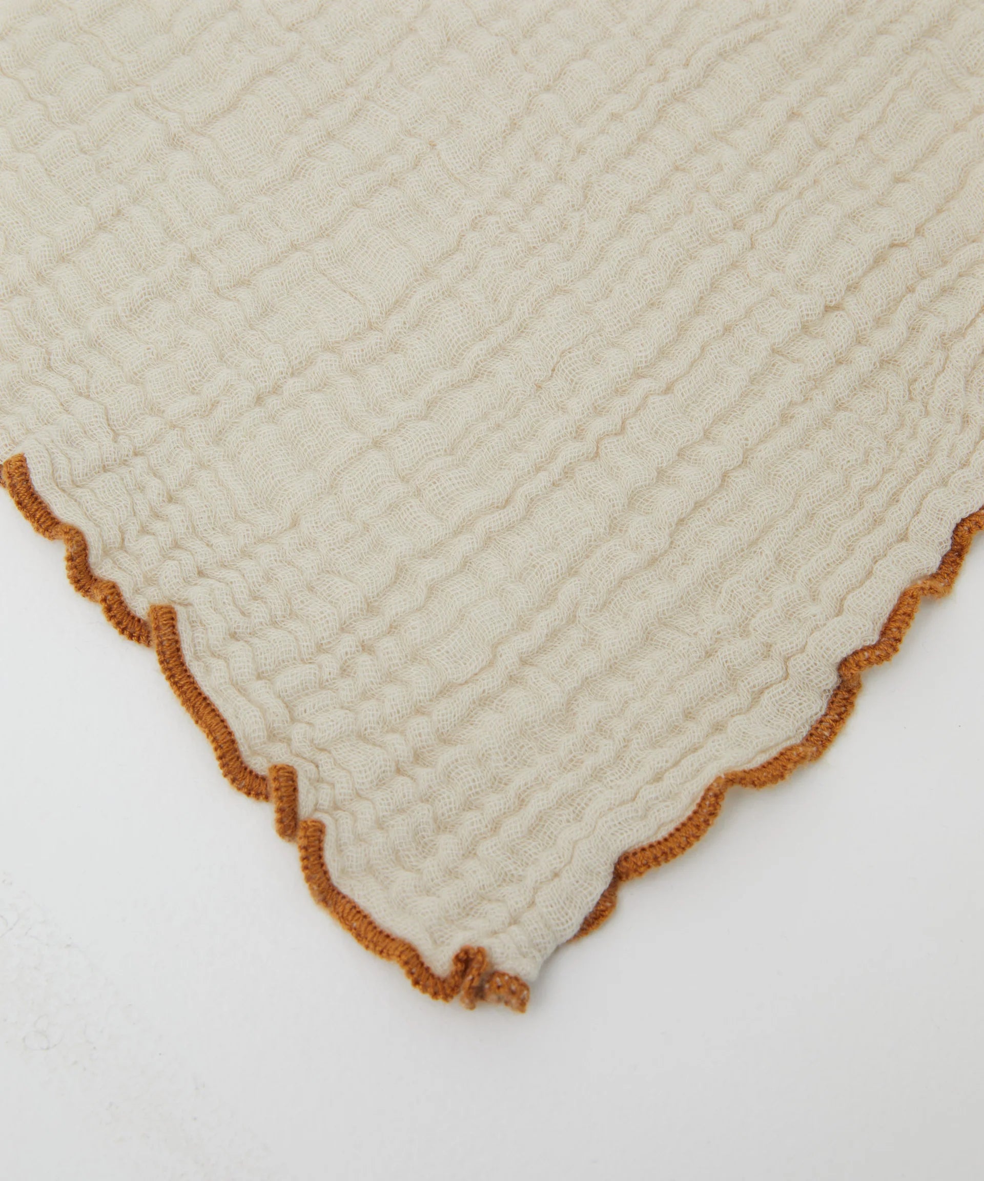 A close-up of PHILÉ's Swaddle Crema from the Cotton Classics Collection showcases a cream-colored, crinkled fabric with brown trim and scalloped edges, crafted from Oeko-Tex Certified Cotton.