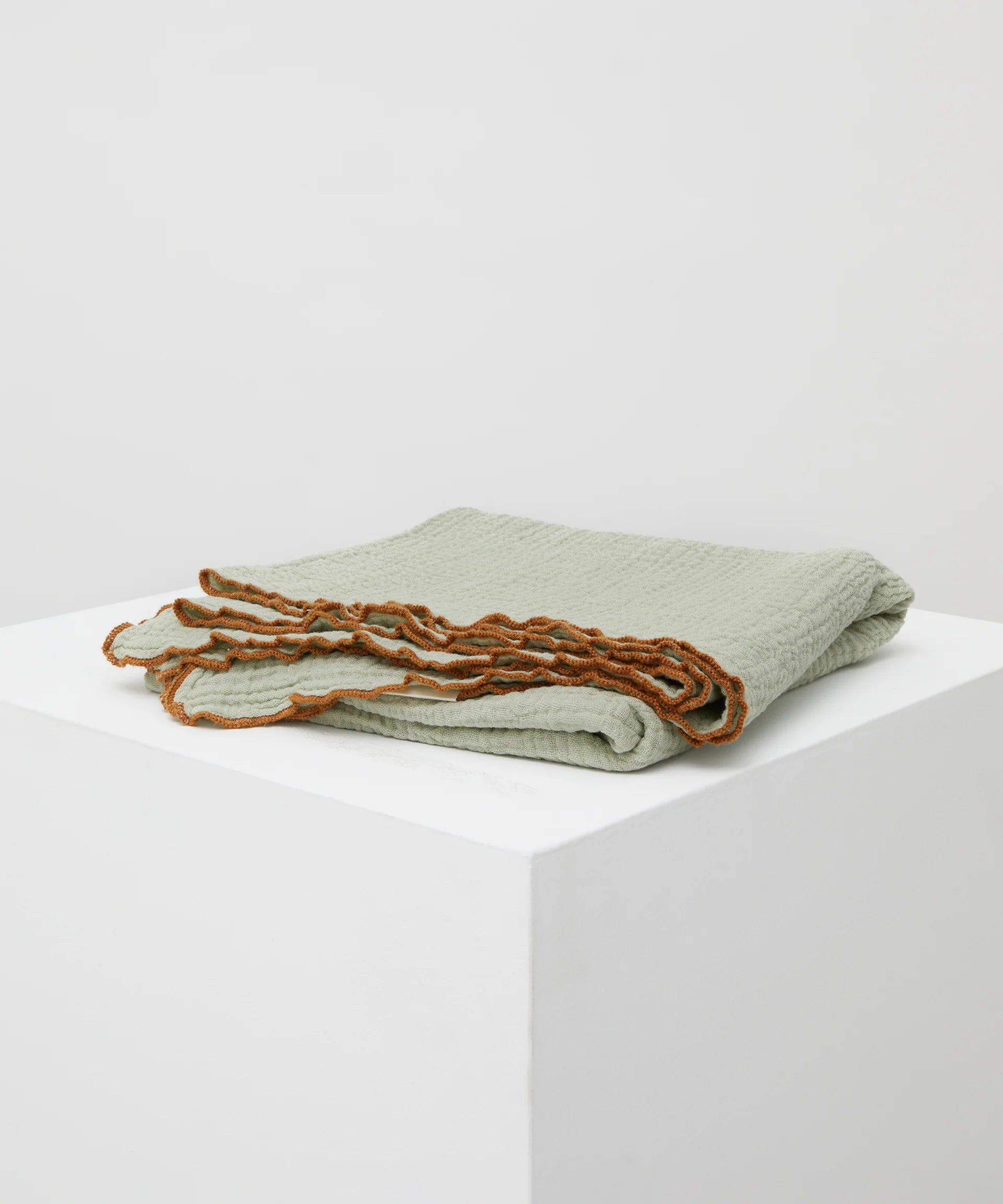 The Swaddle Sage by PHILÉ, a light green muslin wrap with ruffled brown edges, is elegantly displayed on a white pedestal against a plain gray background.