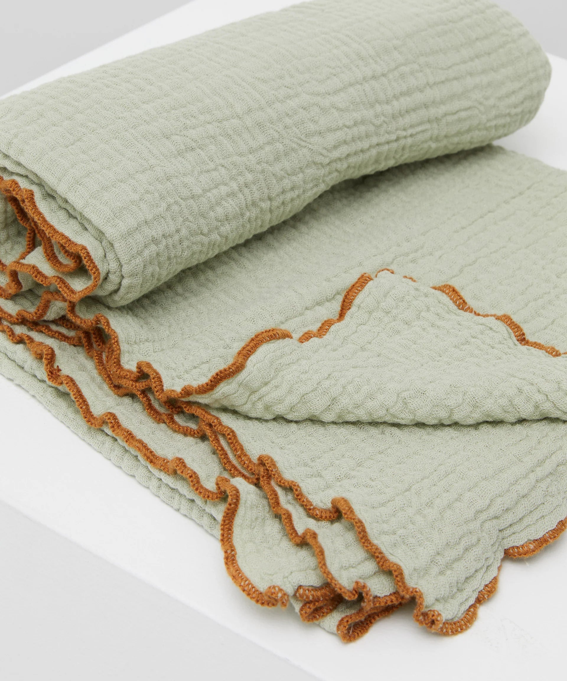 The Swaddle Sage by PHILÉ, a light green blanket made from Oeko-Tex Certified Cotton with a textured waffle pattern and rust-colored ruffled edges, is elegantly placed on a white surface.